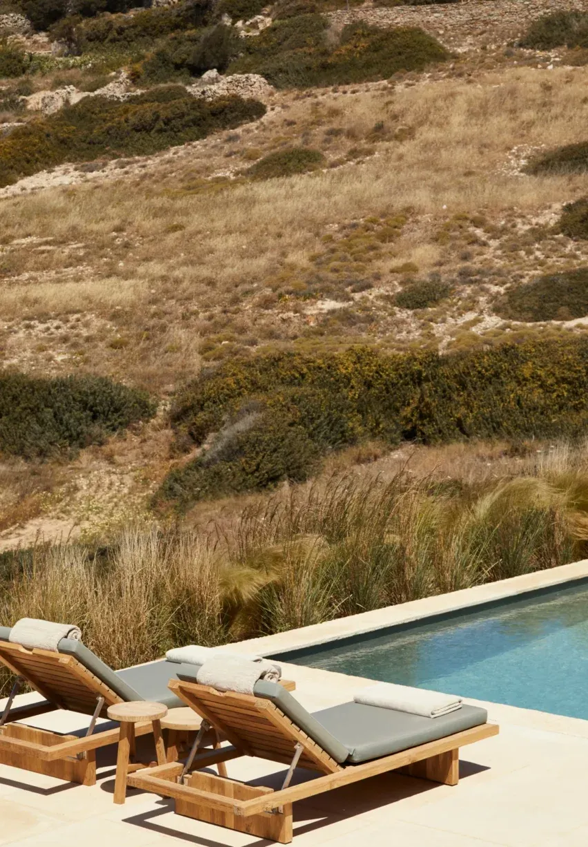 Antiparos, Europe — Teak loungers with cushions beside infinity pool overlooking terraced hillside with native grasses and Mediterranean scrub landscape.
