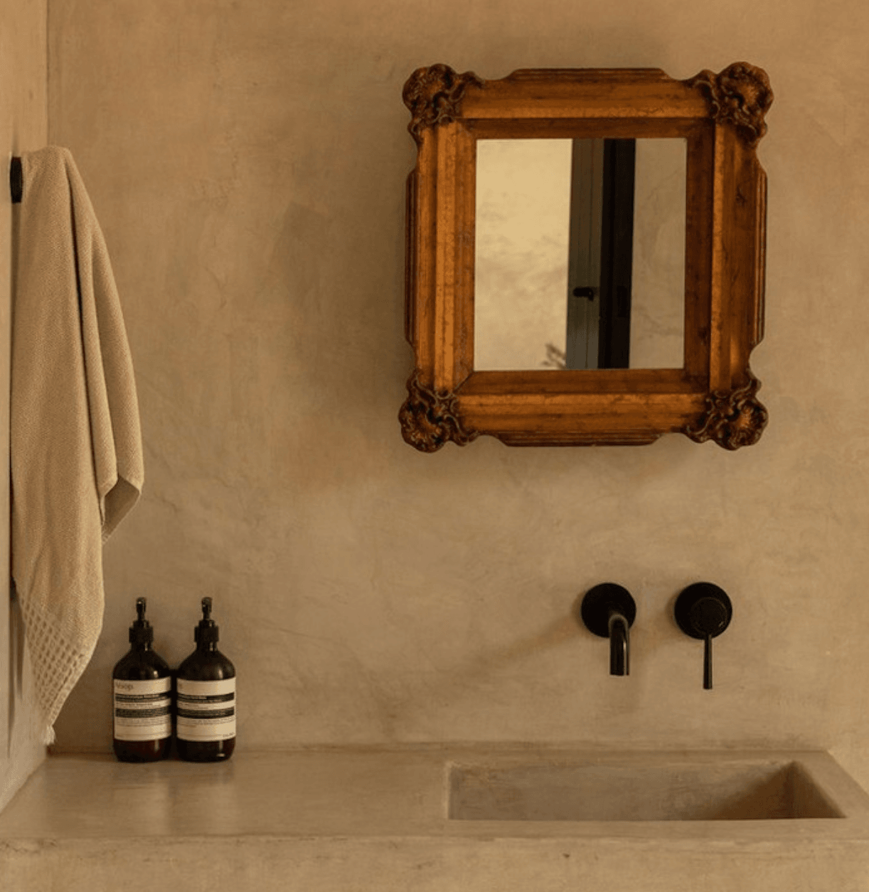 Villa ITO, Mexico — Minimalist bathroom vanity with carved wooden mirror, concrete countertop, black wall-mounted faucet, and dark glass bottles against neutral plaster wall.