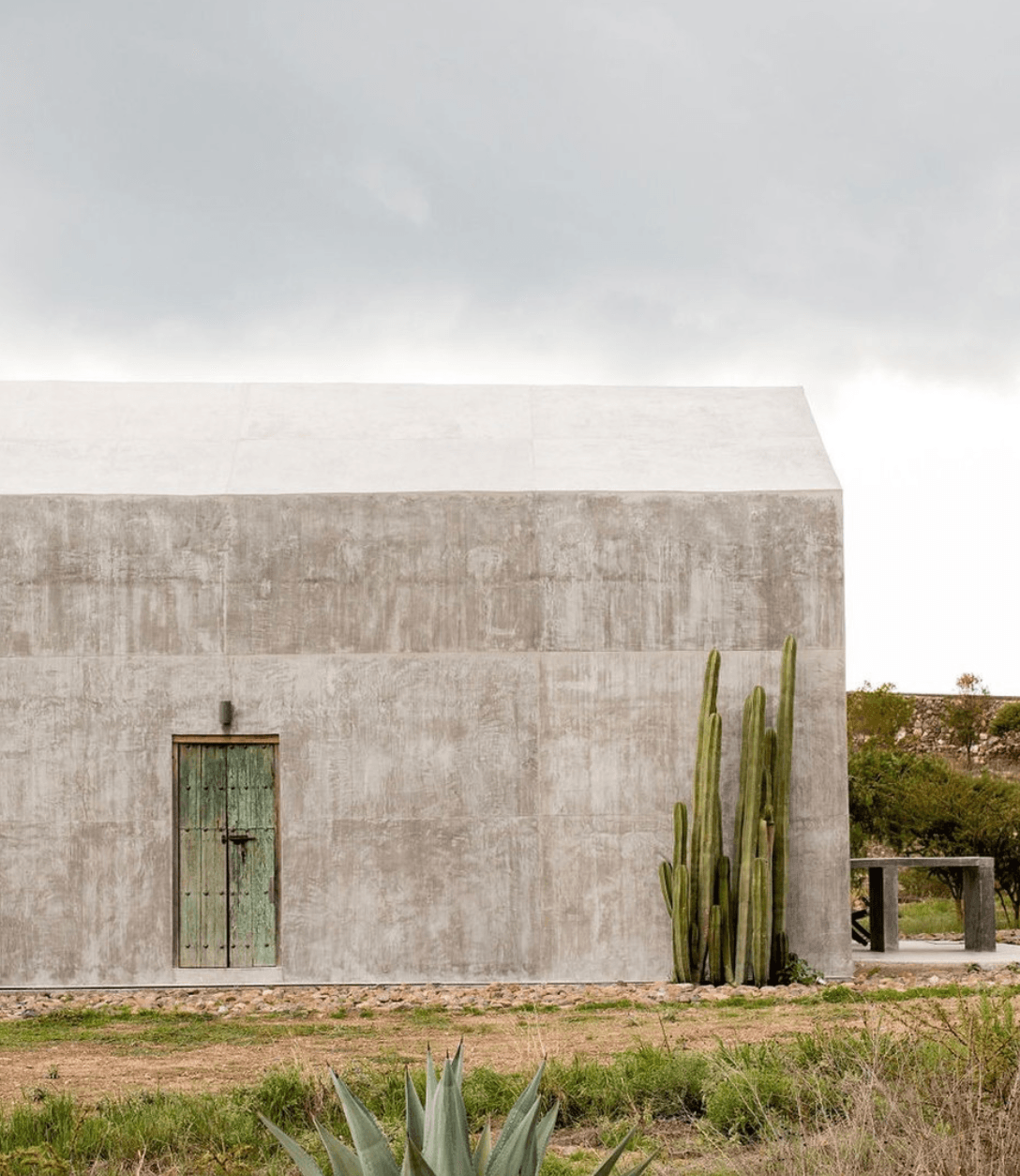 Villa ITO, Mexico — Minimalist concrete structure with weathered facade, tall columnar cacti, and oxidized green door under overcast sky.