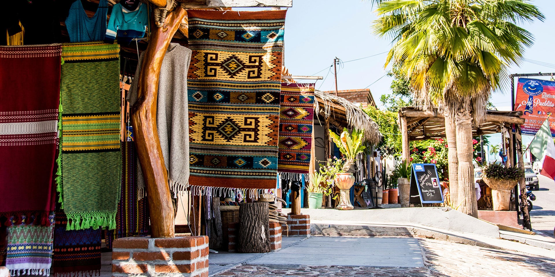 Pueblo Mágico, Mexico — Colorful woven textiles and rustic wooden posts line a sunlit plaza with palm trees and colonial architecture.