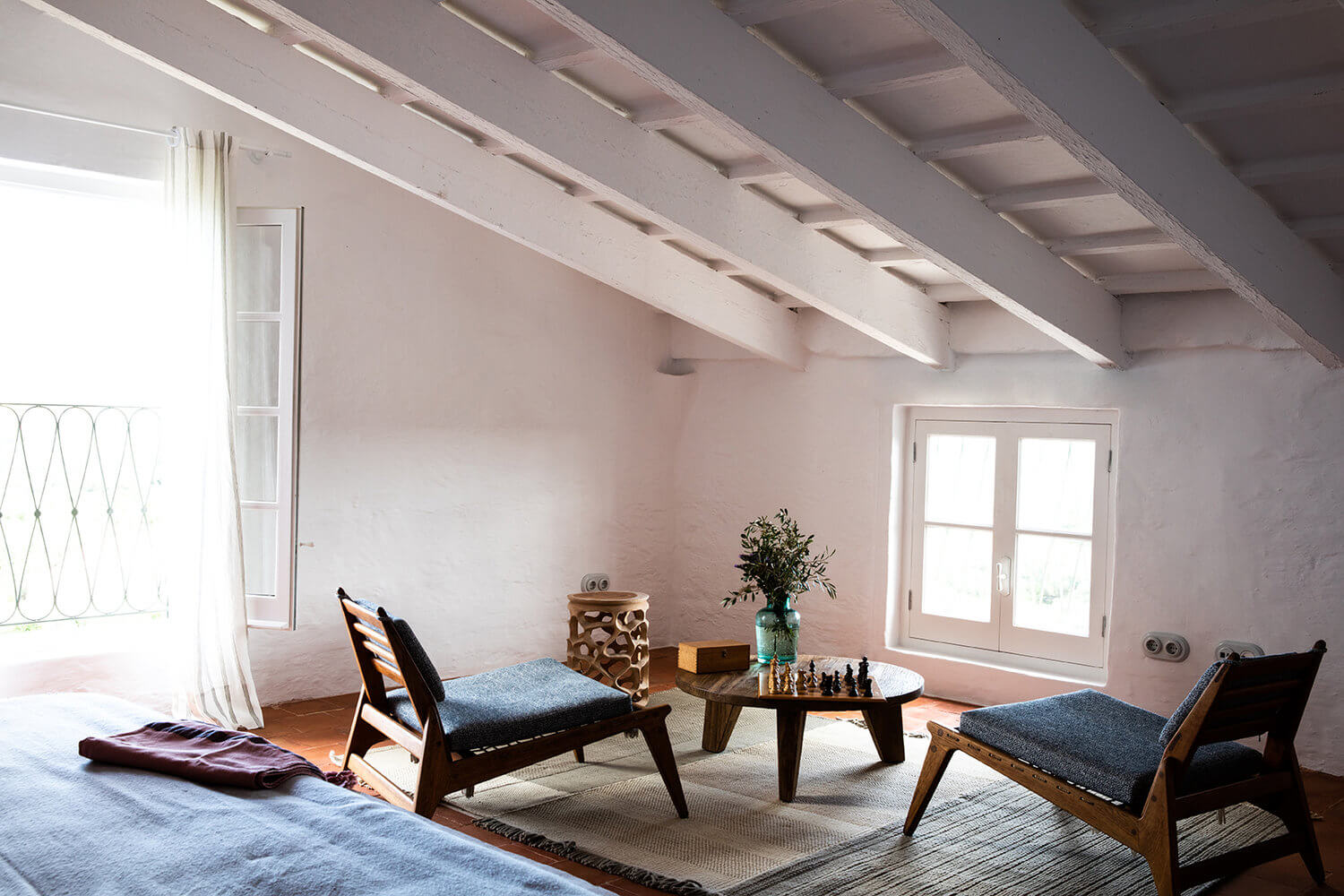 Es Bec d’Aguila — Es Bec d'Aguila, Balearic Islands, Spain — Whitewashed attic room with exposed beams, mid-century wooden chairs, round table with chess set, fresh flowers, and French doors opening to natural light.
