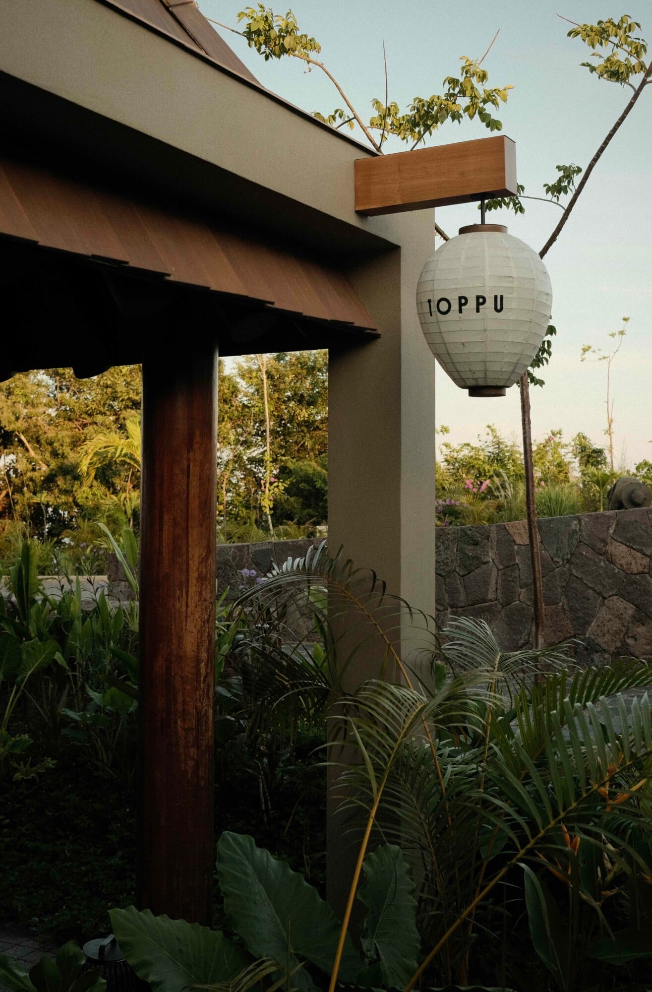 Toppu, Mexico — Modern concrete pavilion with wood accents, spherical paper lantern signage, tropical plants, stone boundary wall at dusk.
