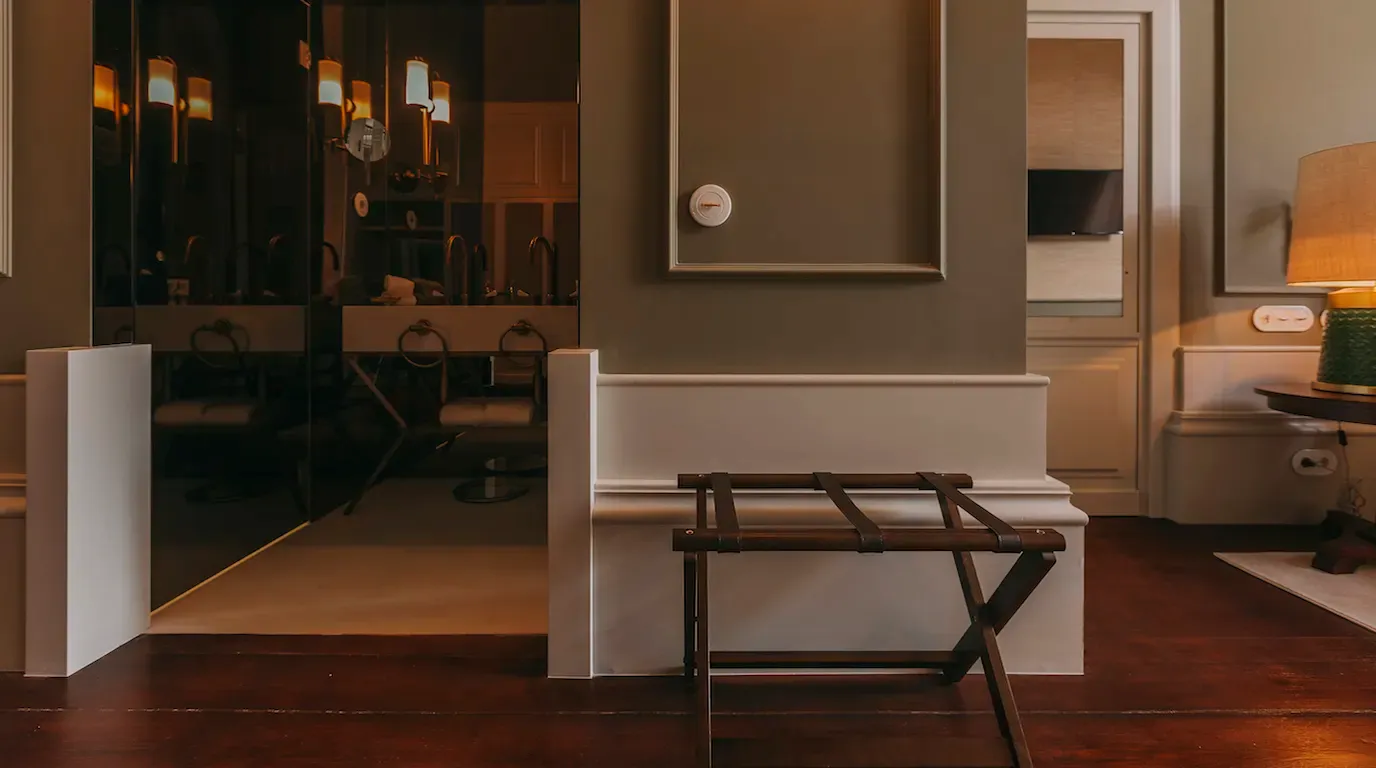 Torel 1884, Porto, Portugal — Minimalist hotel bedroom with white upholstered bench, dark wood luggage rack, warm wood flooring, and bronze wall sconces casting soft ambient light.