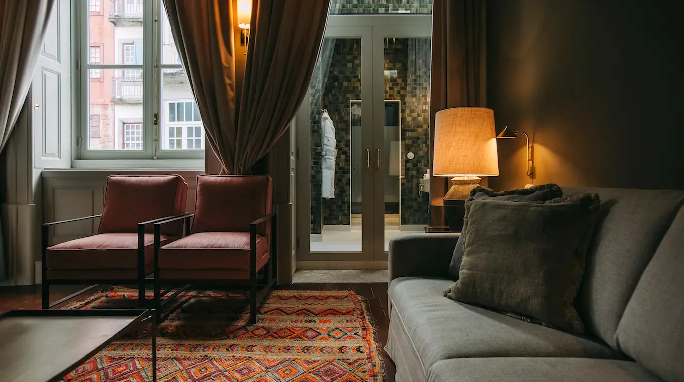 Torel 1884, Portugal — Luxury hotel suite with burgundy mid-century chairs, gray sofa, warm table lamp, geometric patterned rug, floor-to-ceiling windows overlooking historic architecture, and modern bathroom beyond glass doors.