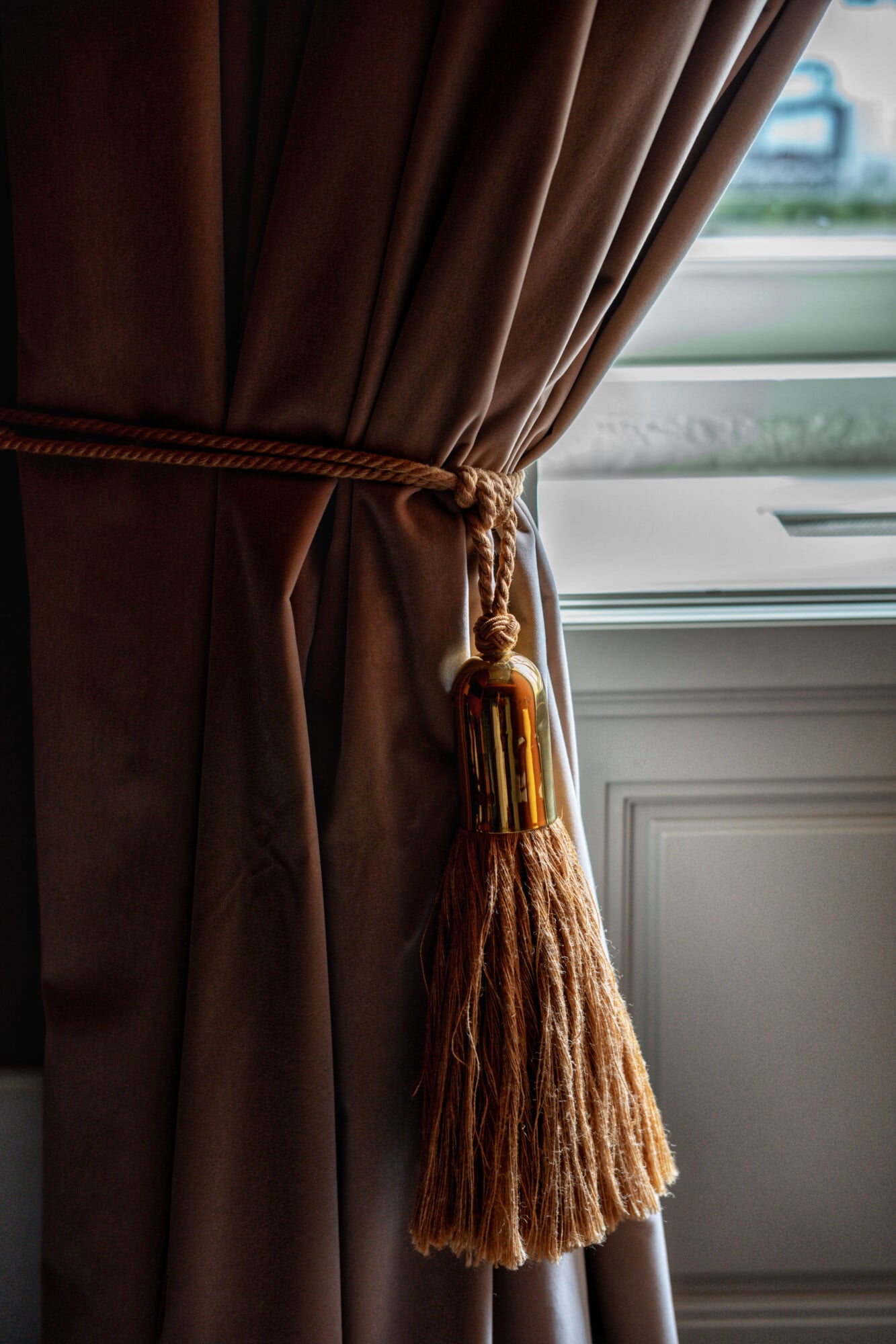 Torel 1884, Portugal — Brown linen curtains with golden tassel tieback beside tall window, natural light illuminating fabric texture and architectural molding.
