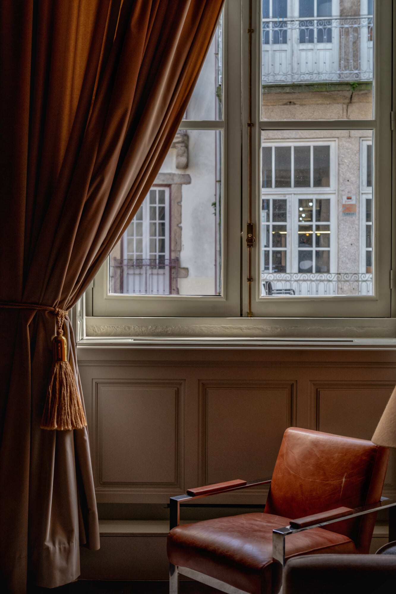 Torel 1884, Portugal — Cognac leather chair by tall sash windows with silk curtains overlooking Lisbon's classical façades and ironwork balconies.