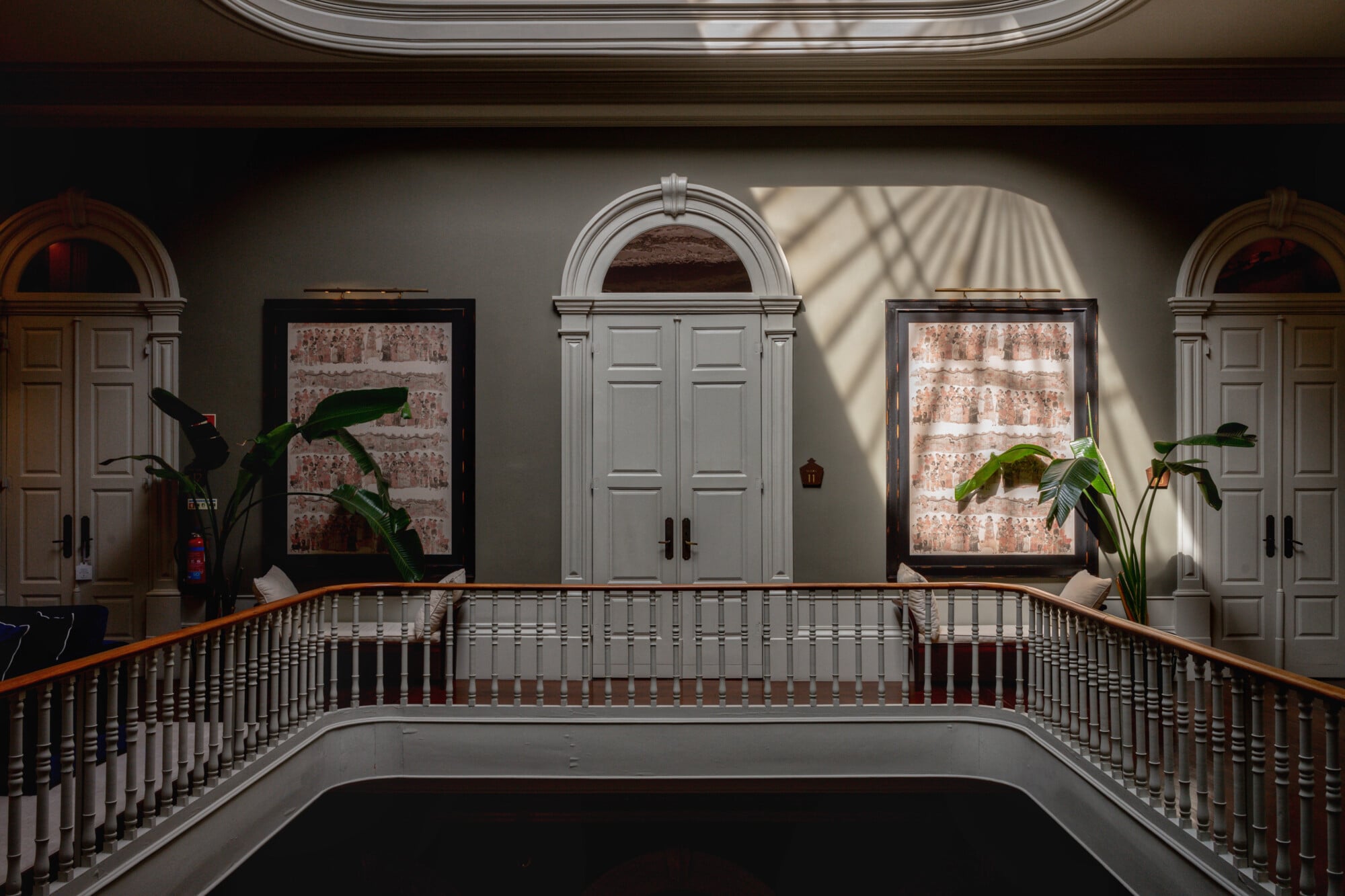 Torel 1884, Portugal — Neoclassical gallery with arched doorways, paneled doors, framed brick artwork, potted plants, and wooden balustrade casting geometric shadows.