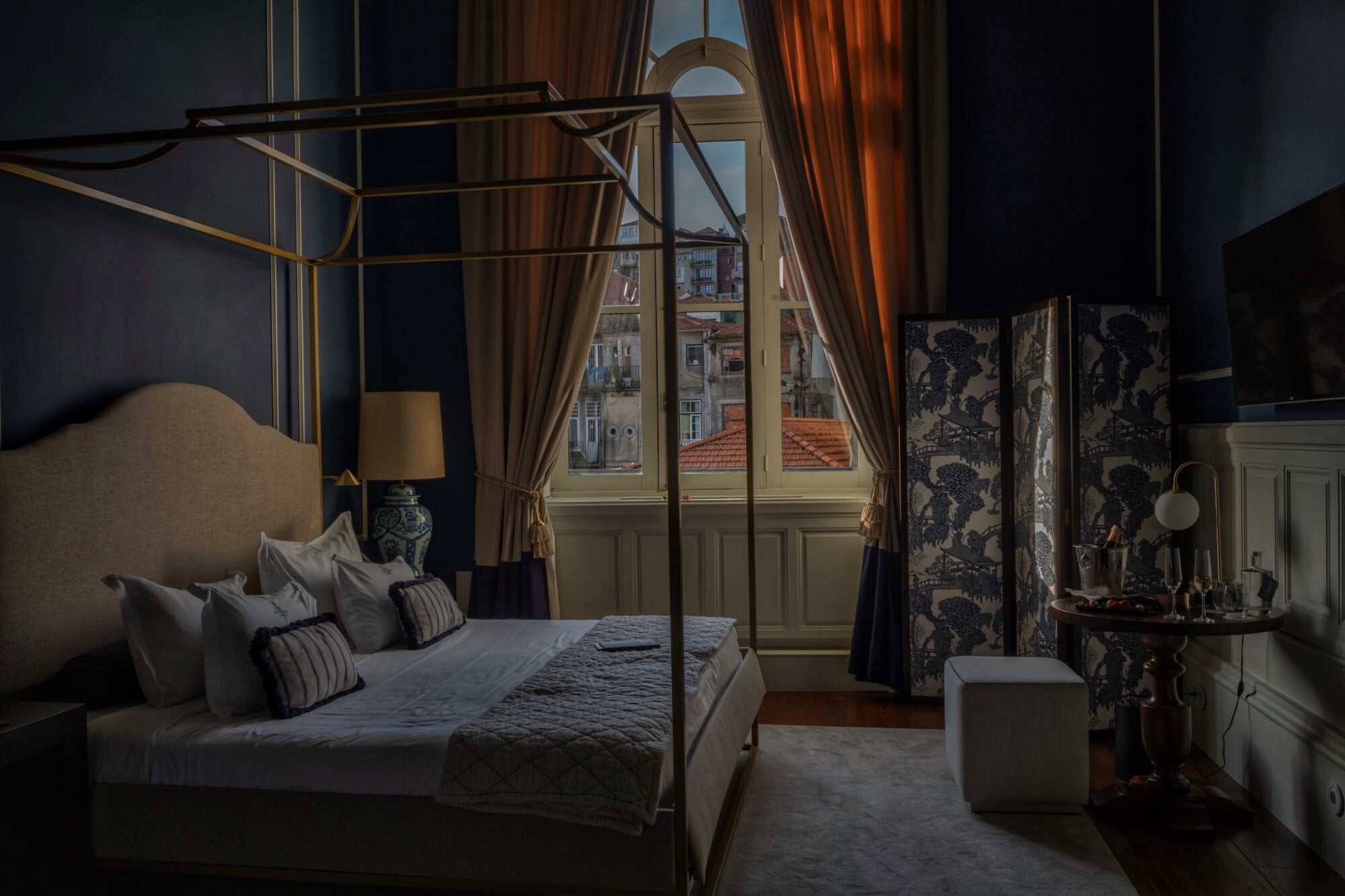 Torel 1884, Lisbon, Portugal — Canopy bed with brass frame in dark bedroom, cream headboard, striped pillows, arched windows with city views, patterned room divider, warm curtains.