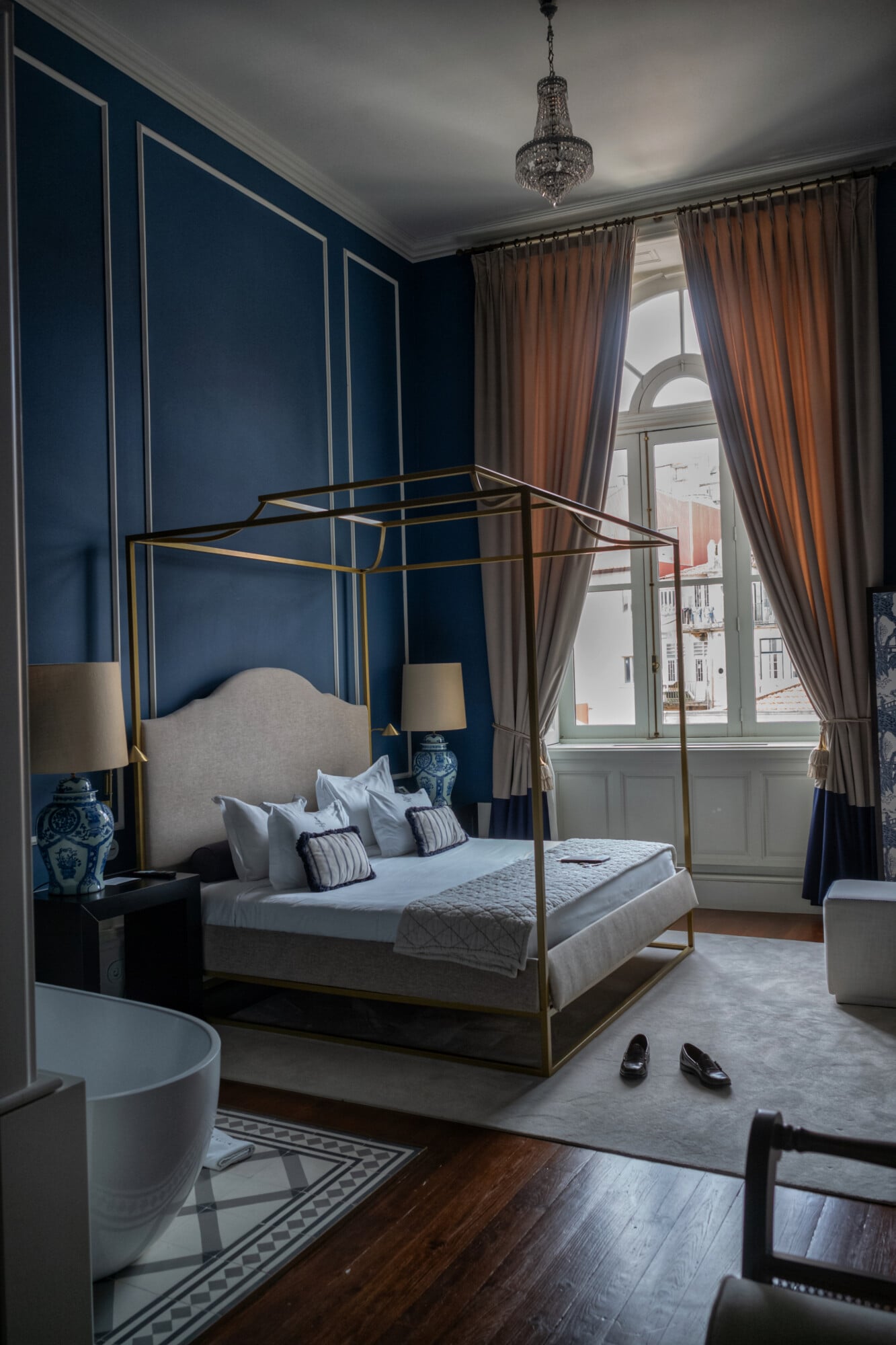 Torel 1884, Portugal — Canopy bed with brass frame against navy paneled walls, arched window with terracotta curtains, crystal chandelier, parquet flooring.