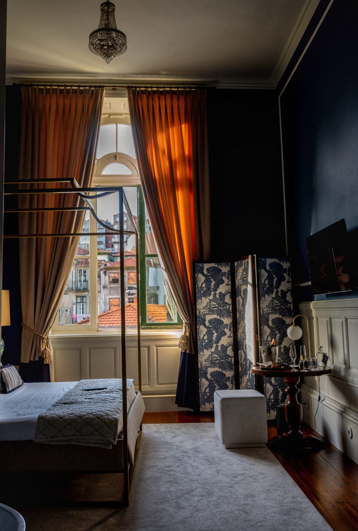Torel 1884, Portugal — Canopy bed in dark-walled bedroom with burnt-orange curtains, arched window overlooking terracotta roofs, ornate chandelier, chinoiserie room divider, brass vanity.