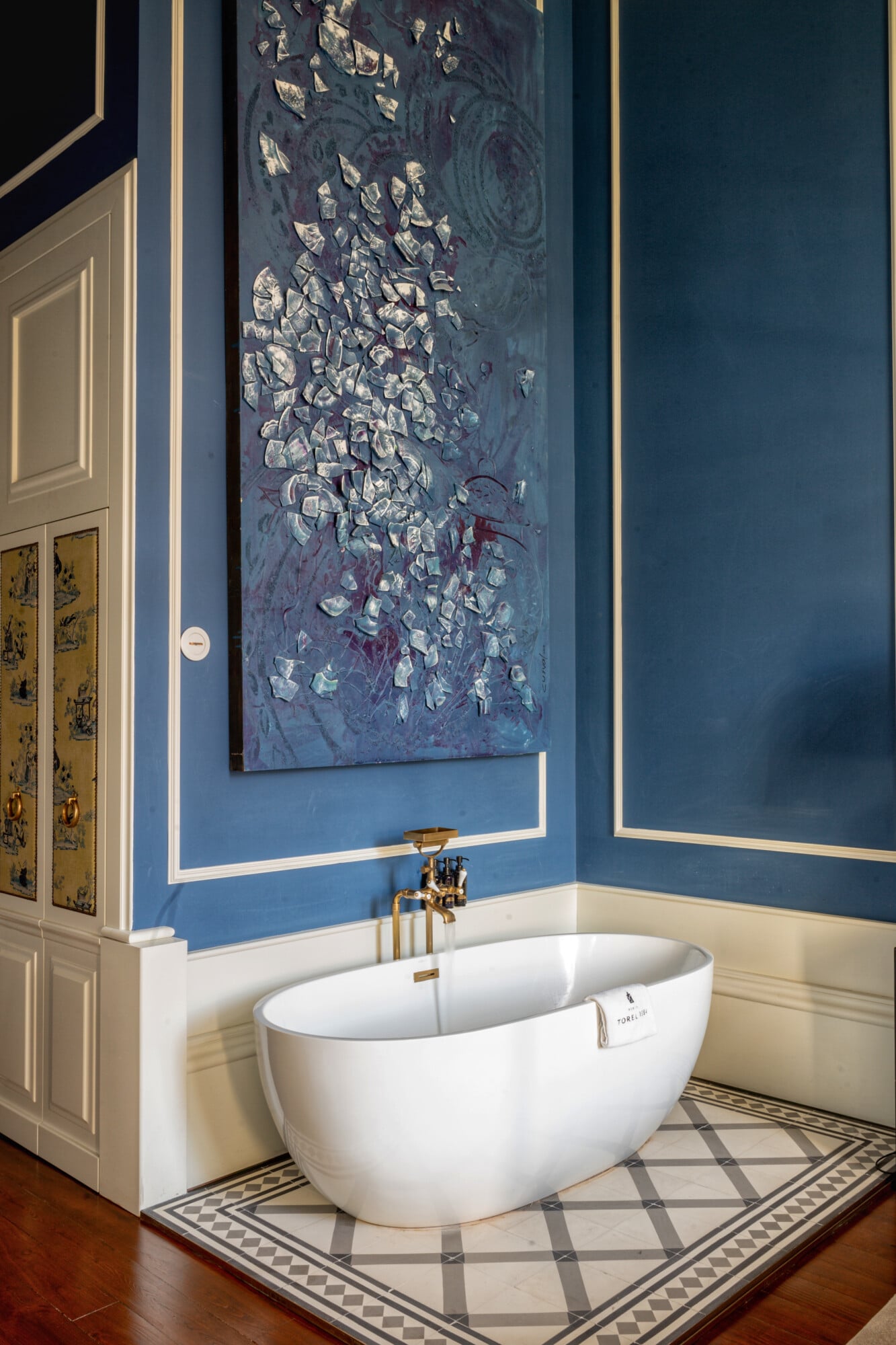 Torel 1884, Portugal — Modern white soaking tub with brass fixtures against deep blue walls and abstract artwork in luxury bathroom with geometric tile floor.