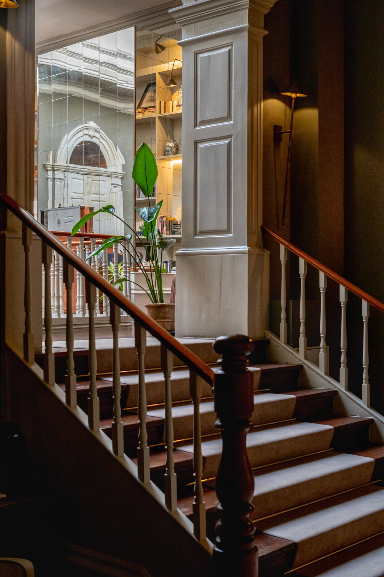 Torel 1884, Portugal — Wooden staircase with brass railings, potted plant, illuminated built-in shelving, arched doorway visible through glass doors, warm ambient lighting.