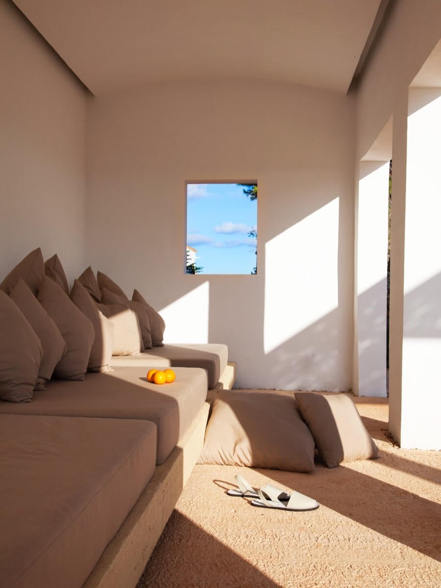 Torralbenc, Balearic Islands — Minimalist coastal bedroom with sand flooring, taupe furnishings, and framed ocean view through window.