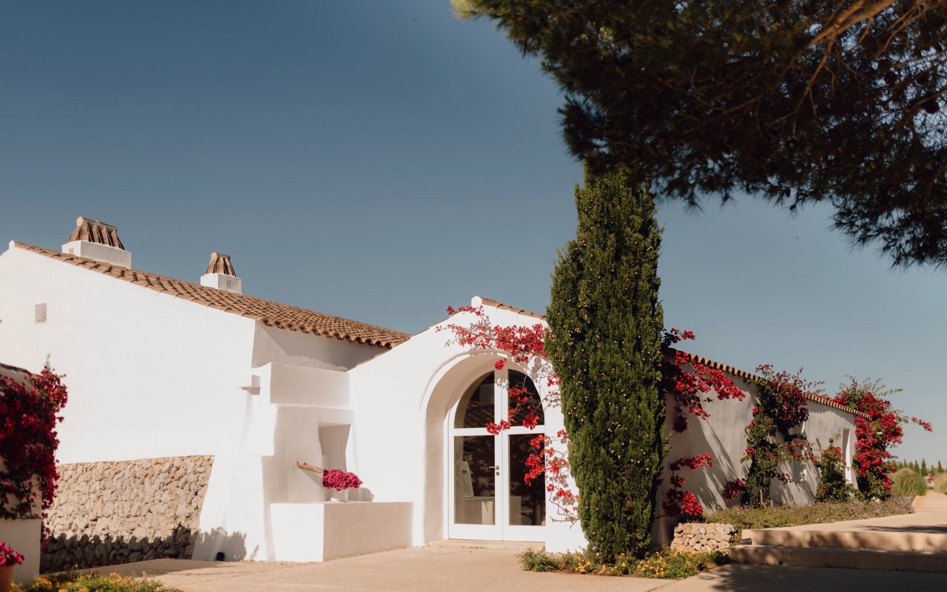 Torralbenc, Balearic Islands — Whitewashed Mediterranean villa with arched doorway, terracotta roof, bougainvillea cascades, cypress trees, minimalist luxury architecture.