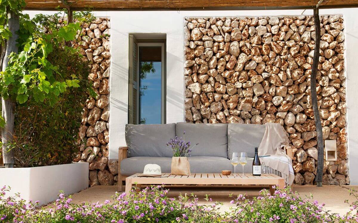 Torralbenc, Balearic Islands — Stone-walled terrace with gray upholstered sofa, wooden table, lavender flowers, and rustic wooden pergola overhead.