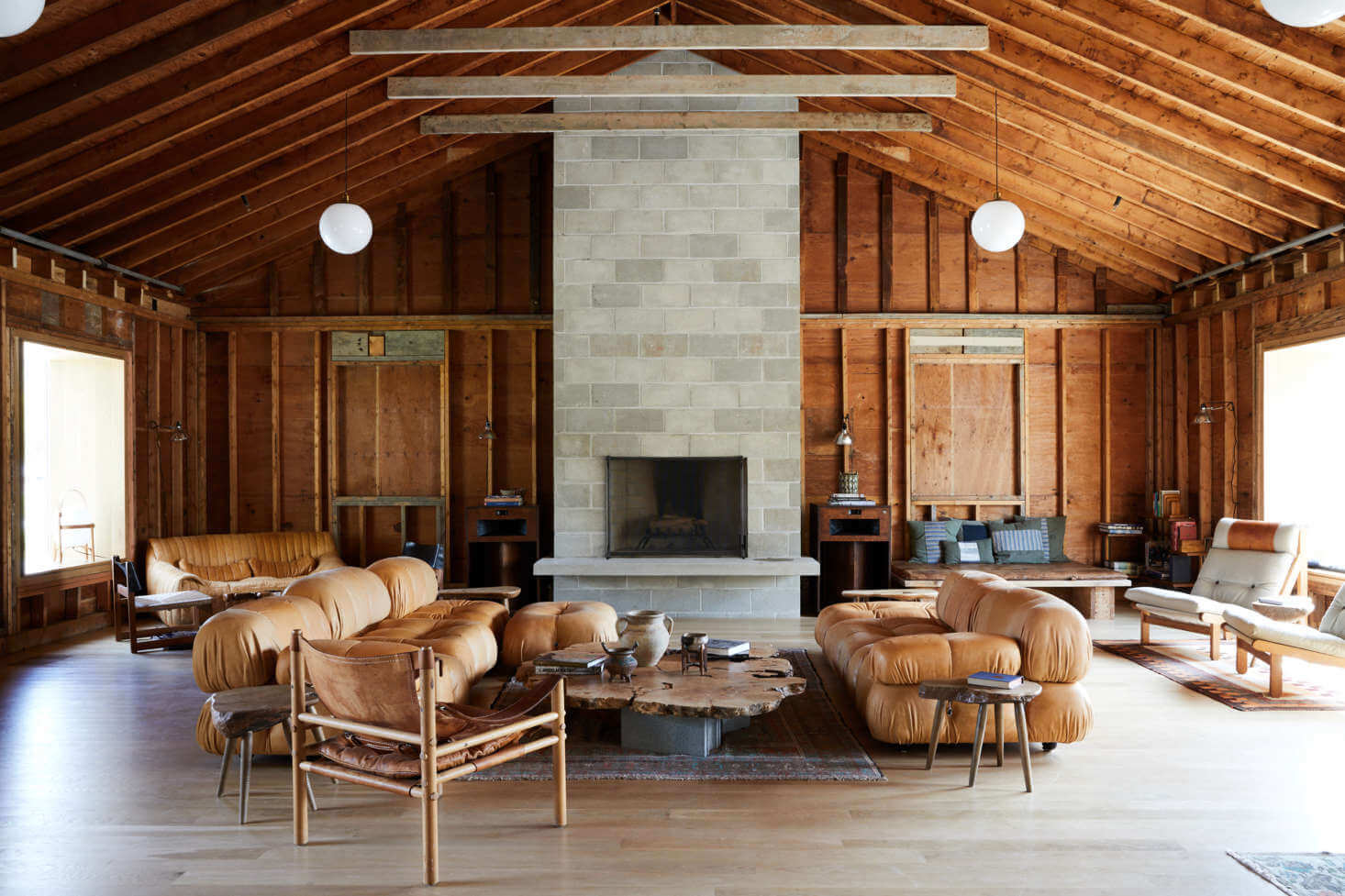 Tourists, North America — Wood-beamed pavilion with central stone fireplace, cognac leather seating, and exposed timber framing creating minimalist luxury interior.