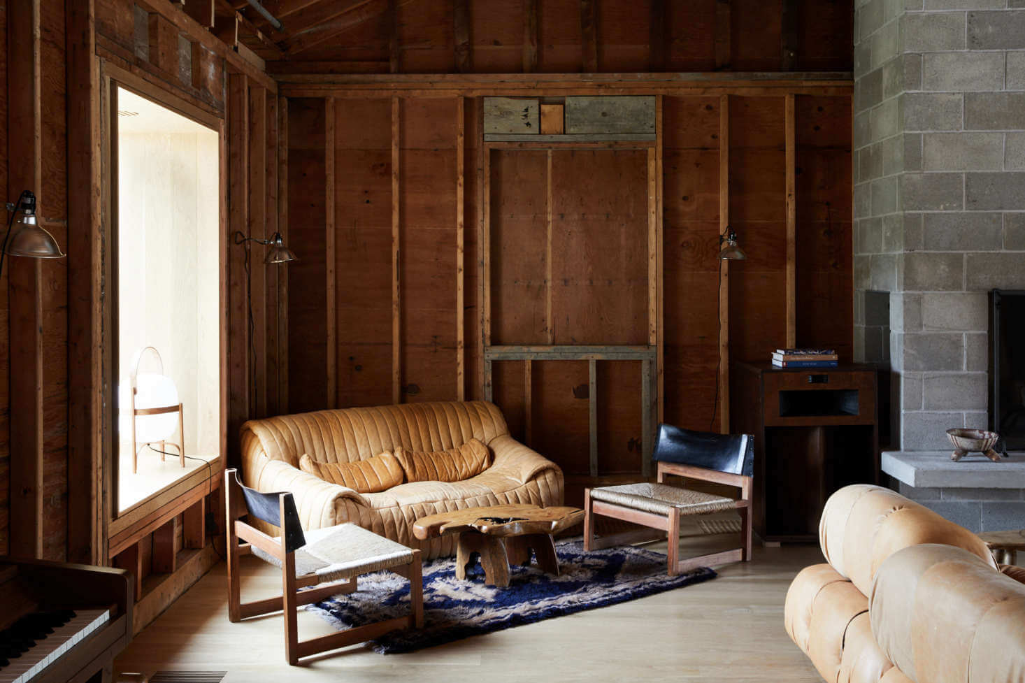 Tourists, North America — Wooden-paneled interior with leather sofa, midcentury furniture, concrete fireplace, and natural light from large window.