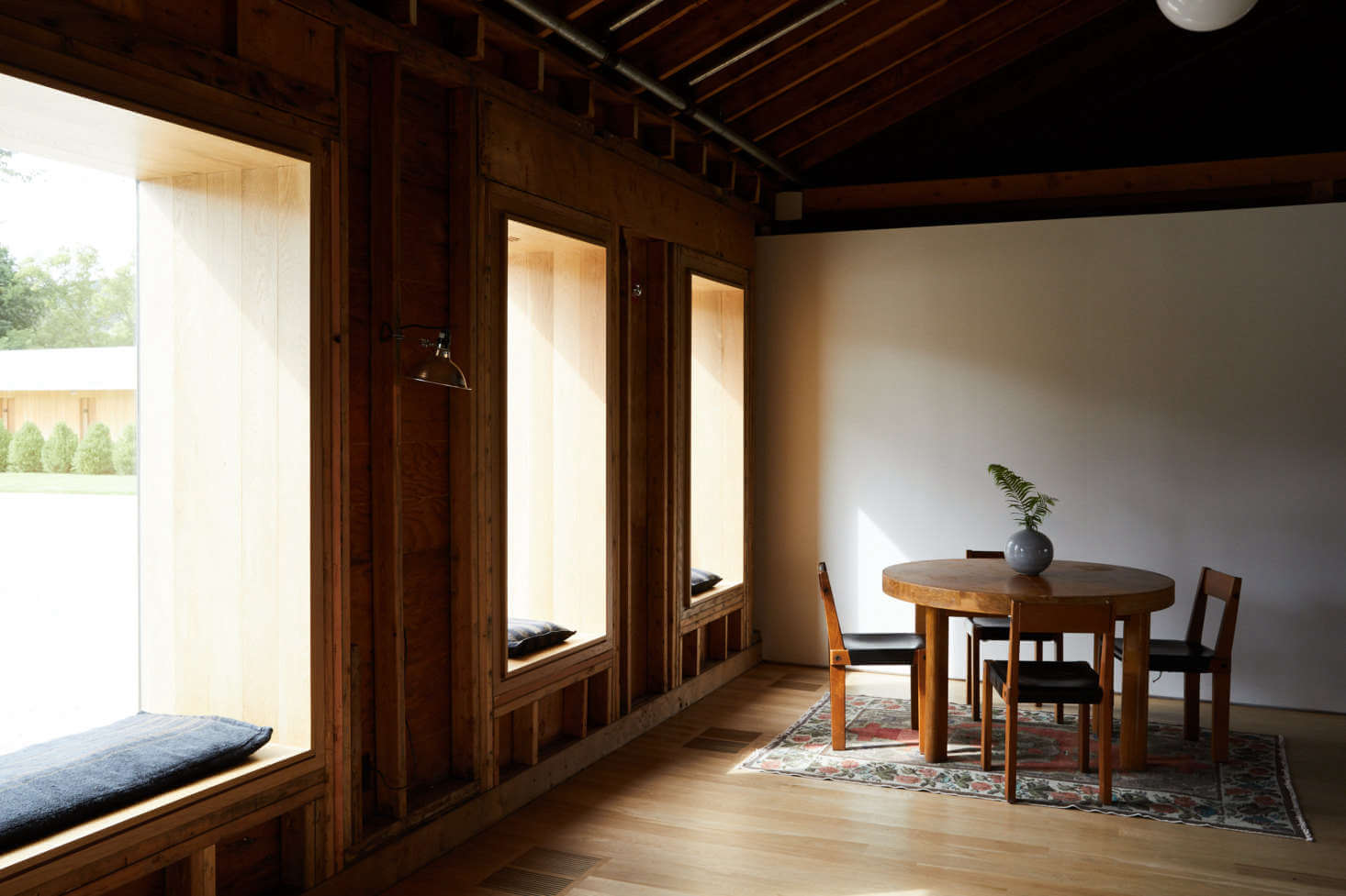 Tourists, North America — Minimalist dining room with dark wood beams, natural light through floor-to-ceiling windows, round wooden table, and Japanese-inspired architectural details.