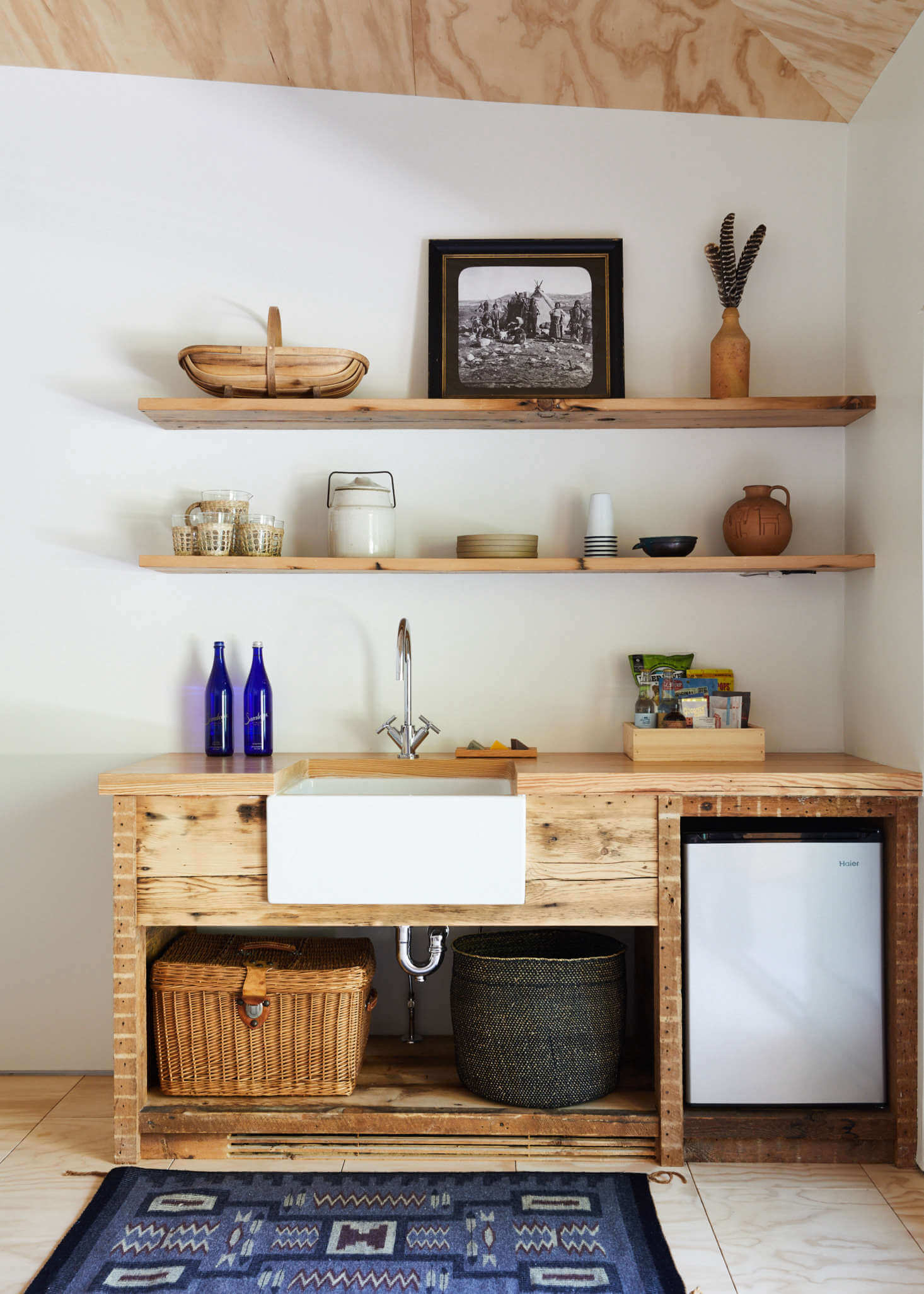Tourists, North America — Reclaimed wood kitchenette with white farmhouse sink, open shelving, woven storage baskets, and minimalist styling against white walls.
