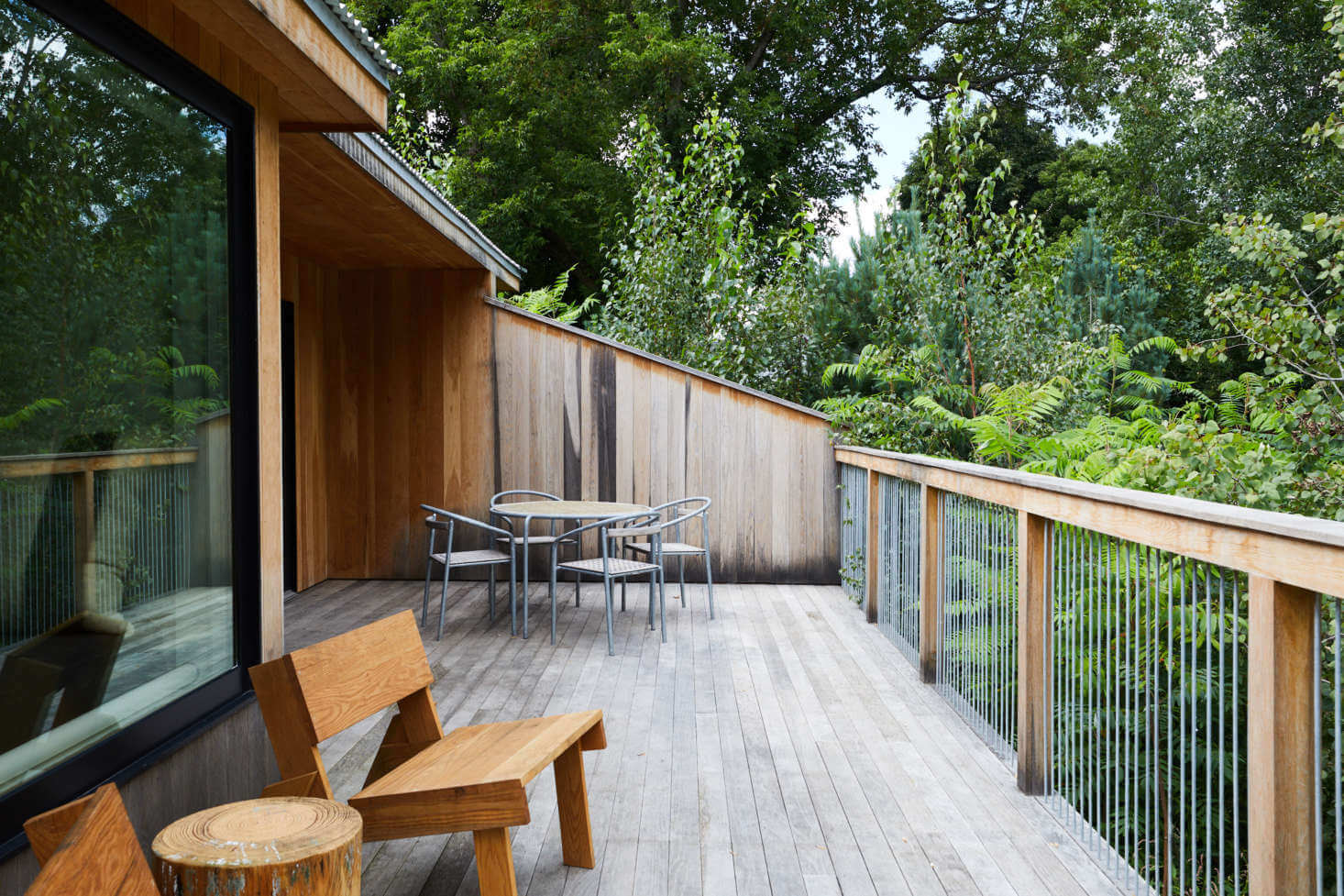 Tourists, North America — Modern wooden deck with metal railings overlooking forest, featuring natural wood furniture and covered pavilion with floor-to-ceiling glazing.