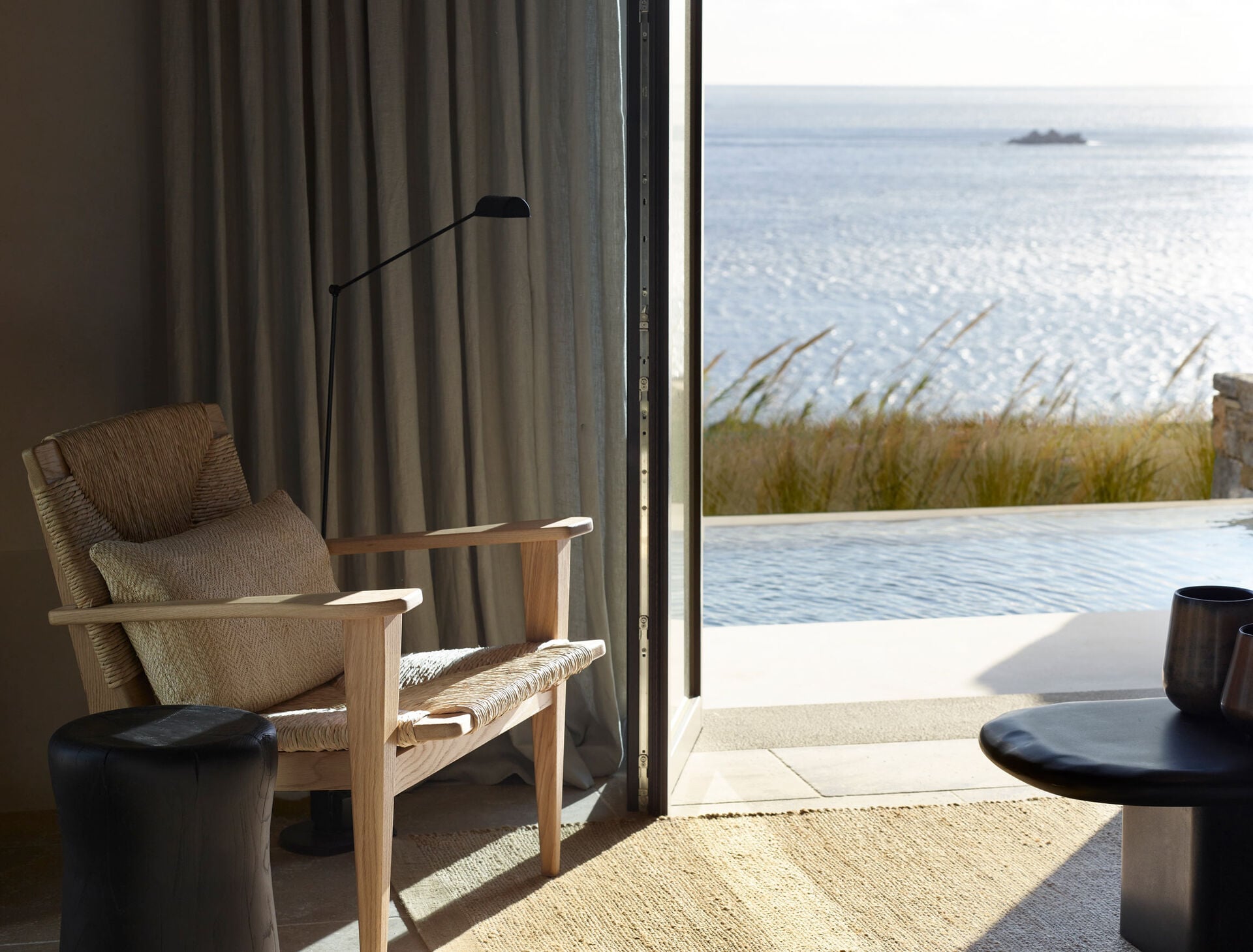 Kalesma, Europe — Minimalist coastal bedroom with wooden armchair, linen cushions, floor-to-ceiling curtains, infinity pool, and sea view with native grasses.