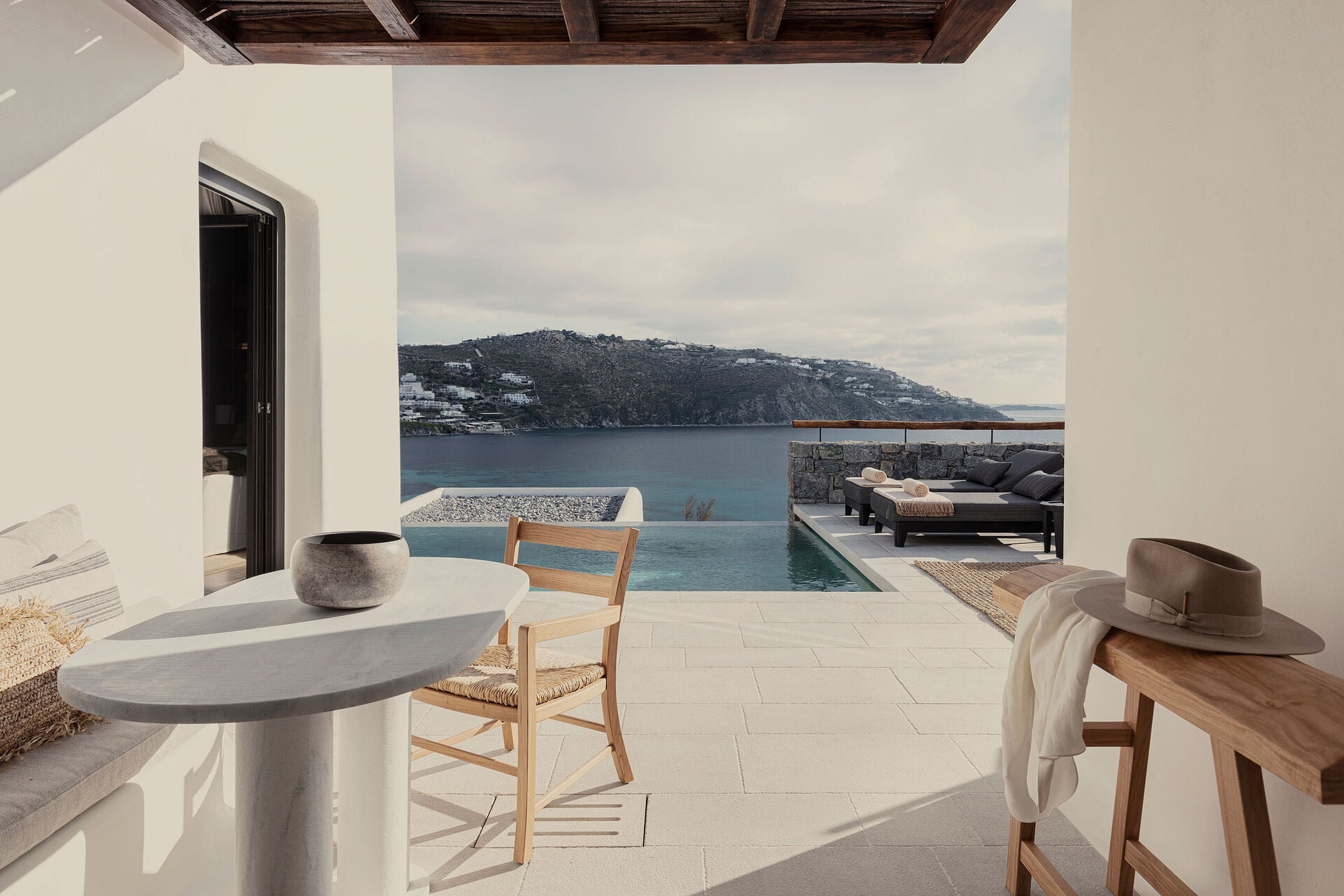 Kalesma, Europe — Modernist clifftop terrace with infinity pool, wooden furniture, stone accents, and sweeping Mediterranean sea views beneath overcast sky.