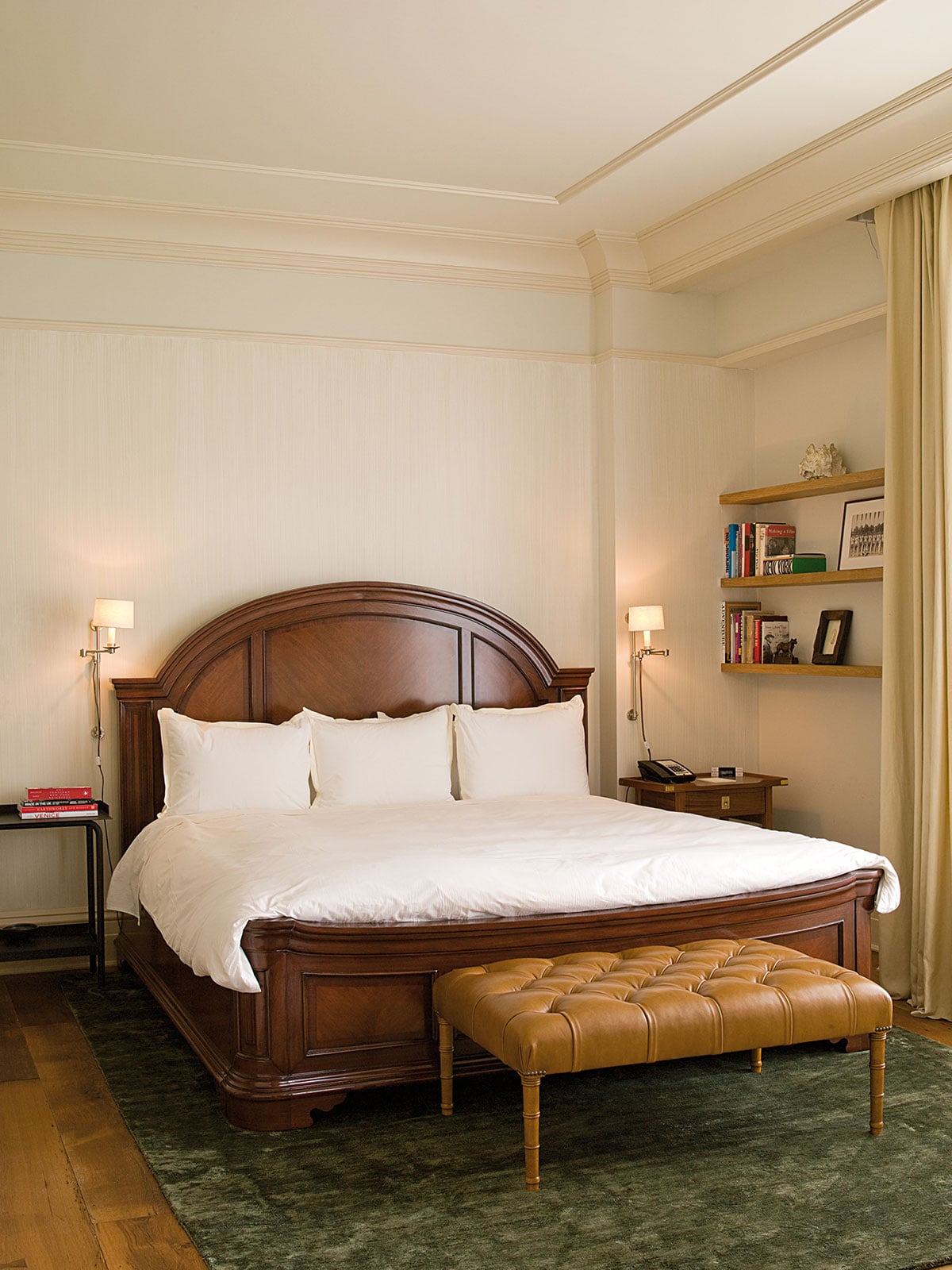 The Greenwich Hotel, New York, USA — Bedroom with curved wood headboard, white linens, leather bench, floating shelves, brass sconces, cream walls, and warm ambient lighting.