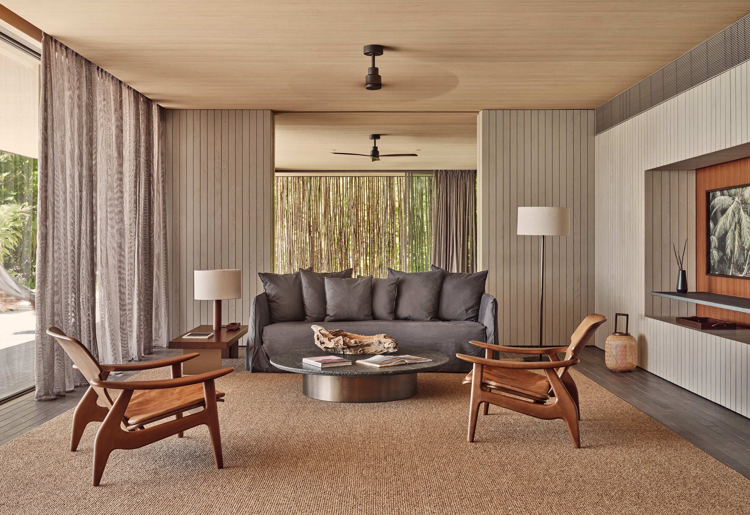 Patina Maldives, Asia — Mid-century modern villa lounge with teak chairs, charcoal sofa, vertical wood paneling, natural light filtering through floor-to-ceiling curtains, and tropical garden views.