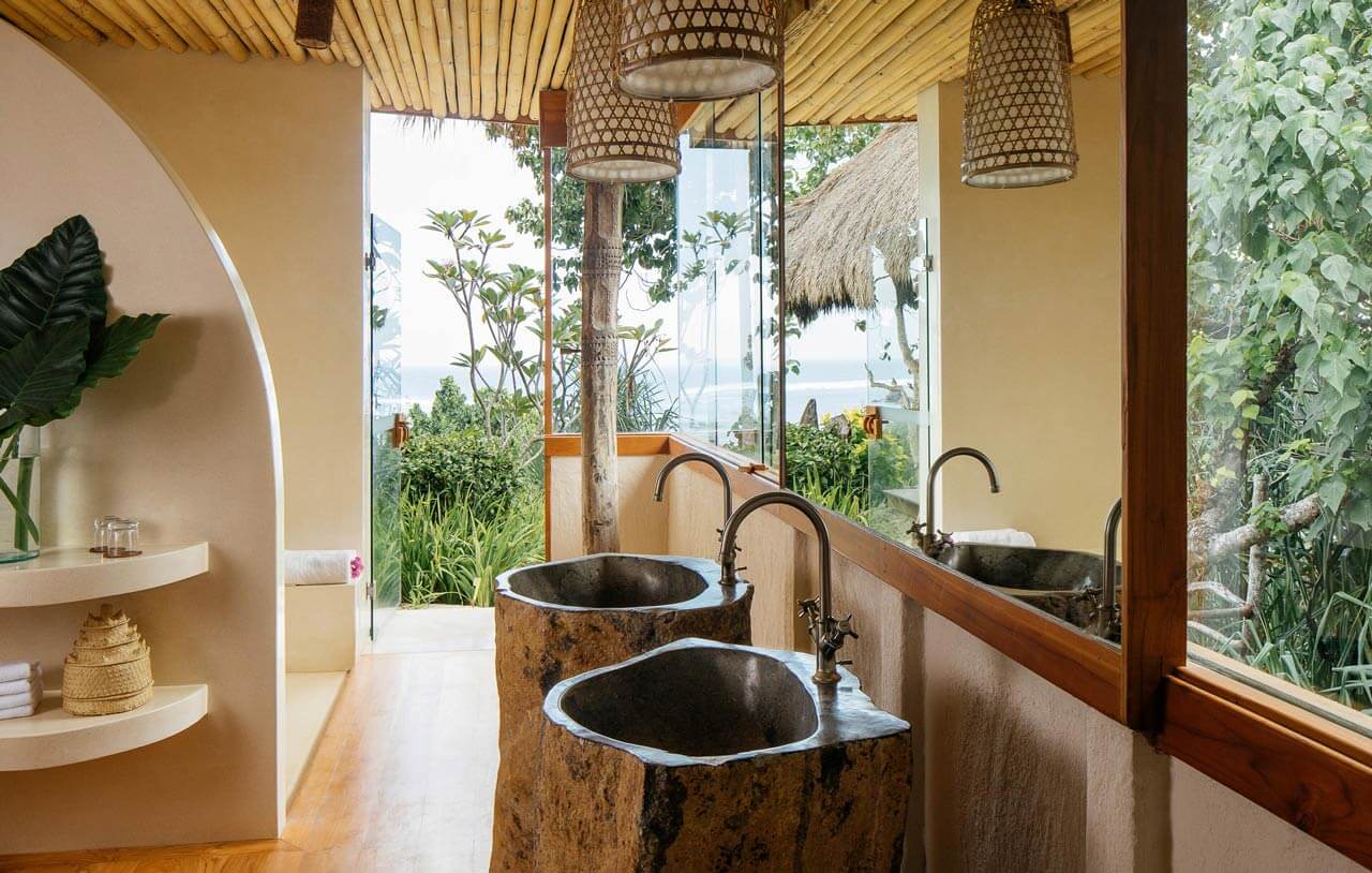NIHI Sumba, Asia — Tropical bathroom with volcanic stone vessel sinks, wooden ceiling details, woven pendant lights, garden views, and ocean horizon beyond glass doors.