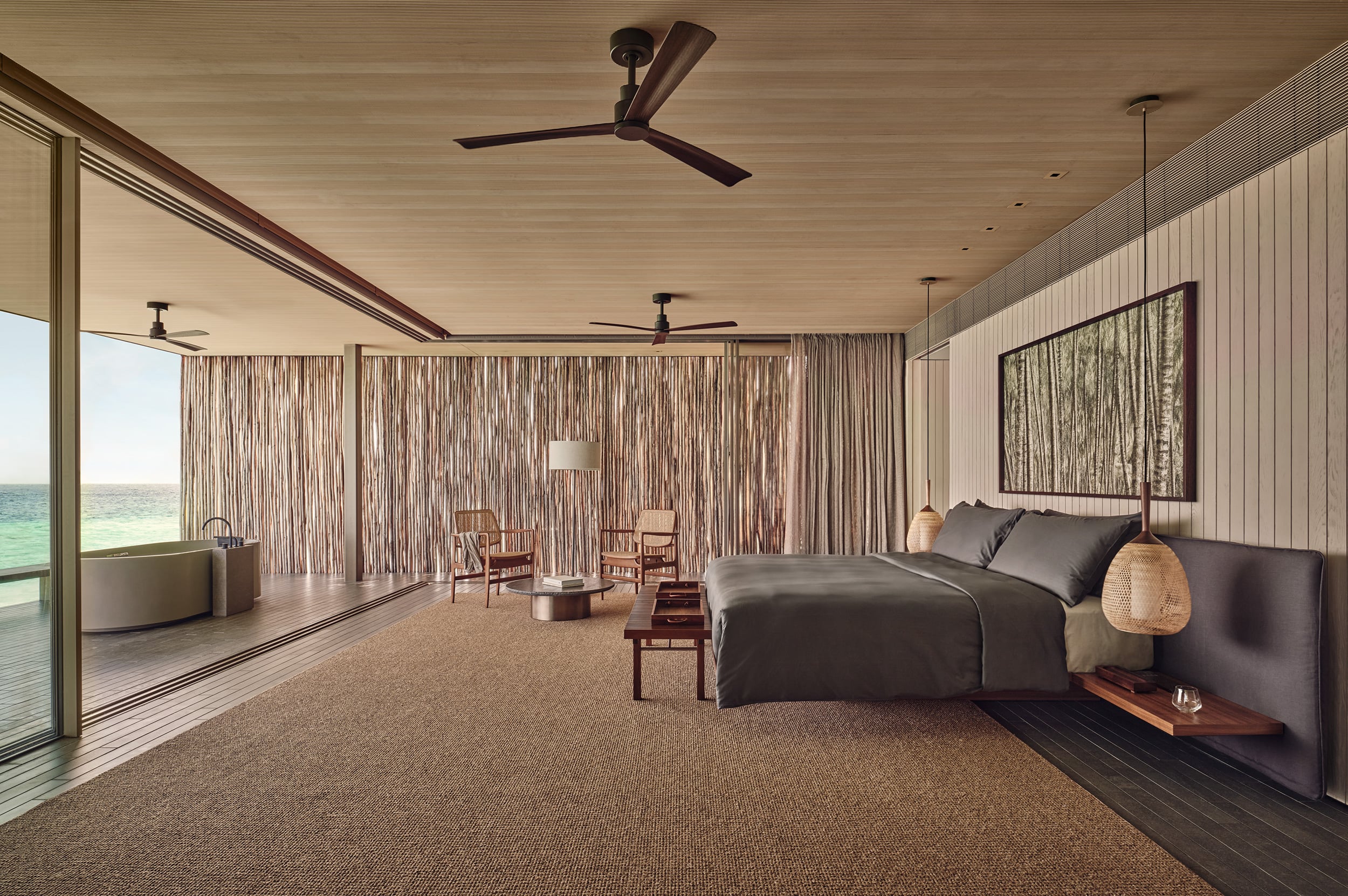 Patina Maldives, Maldives — Overwater villa bedroom with ocean views, woven curtains, dark bed, timber ceiling, and teak furnishings.
