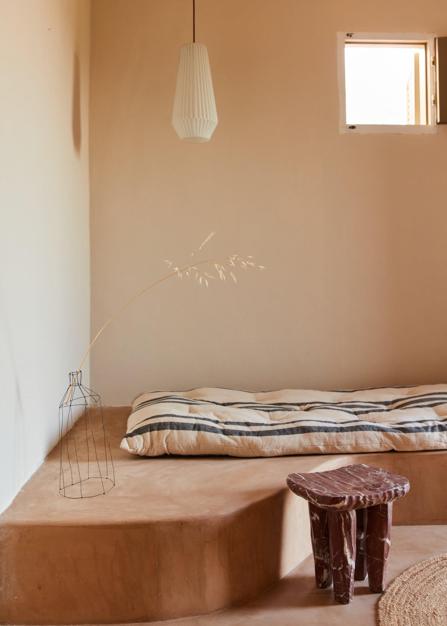 Parco Dei Sesi, Europe — Minimalist bedroom with terracotta walls, pleated pendant light, striped bedding, wire vase with dried grasses, and sculptural wooden stool.