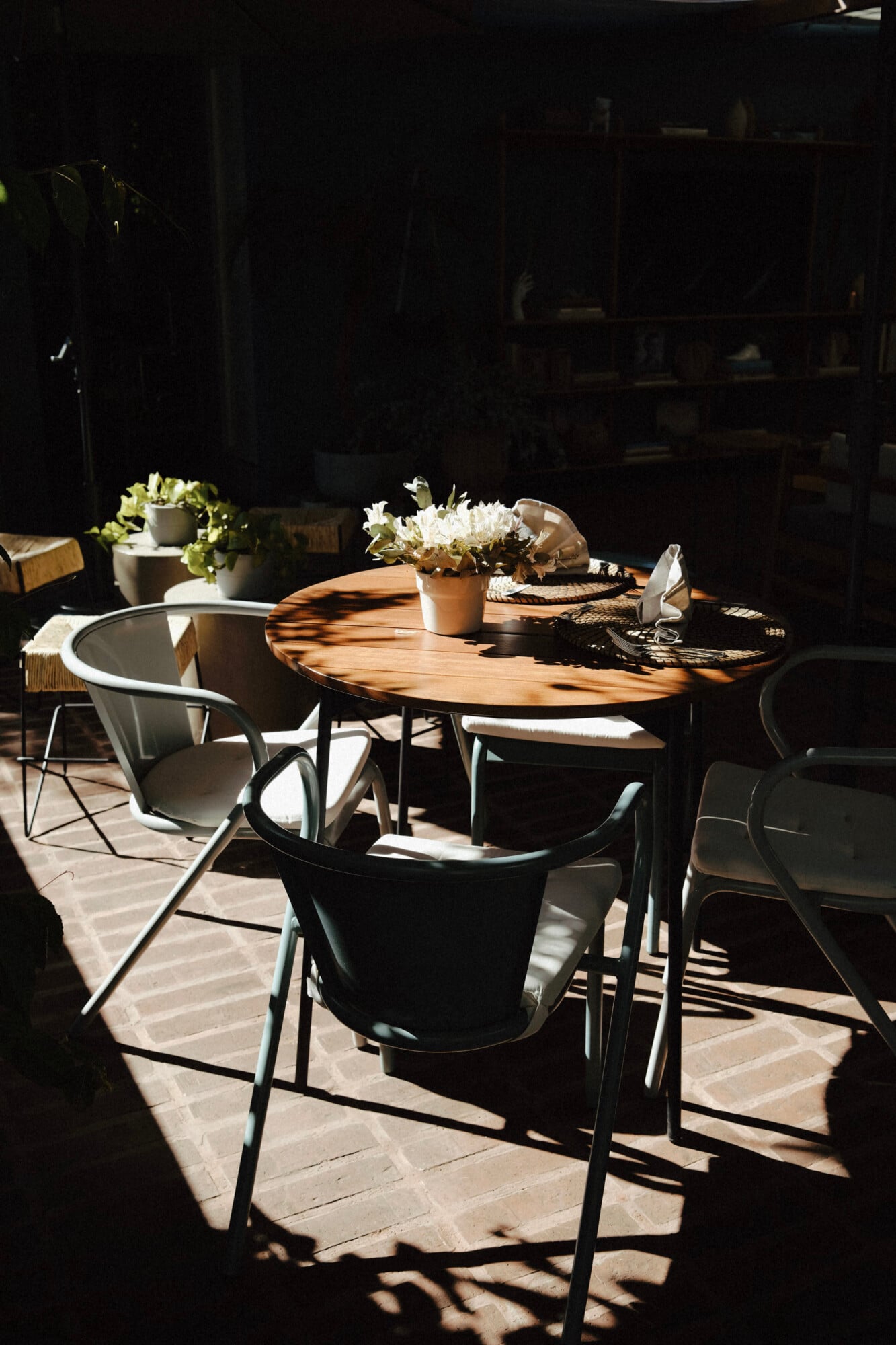 UMA Casa, Mexico — Wooden round dining table with black modern chairs on stone patio, white flowers in terracotta planters, dappled sunlight casting shadows, minimalist luxury outdoor setting.