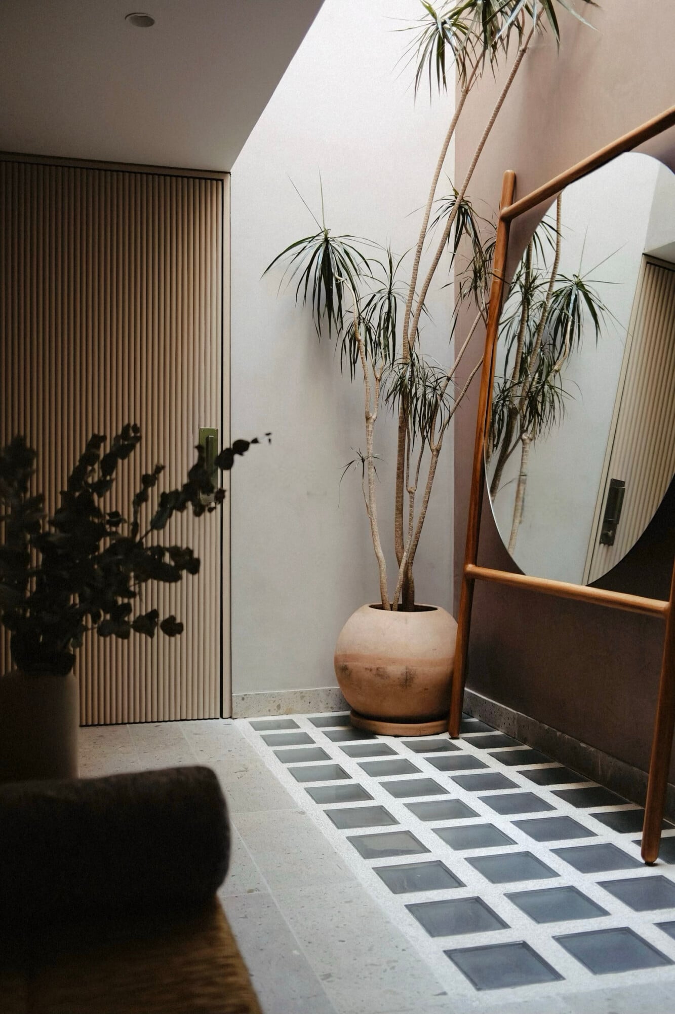 UMA Casa, Mexico — Minimalist entryway with potted dracaena plant in terracotta vessel, wooden-framed mirror, corrugated door, geometric tile floor, and natural light from skylight.