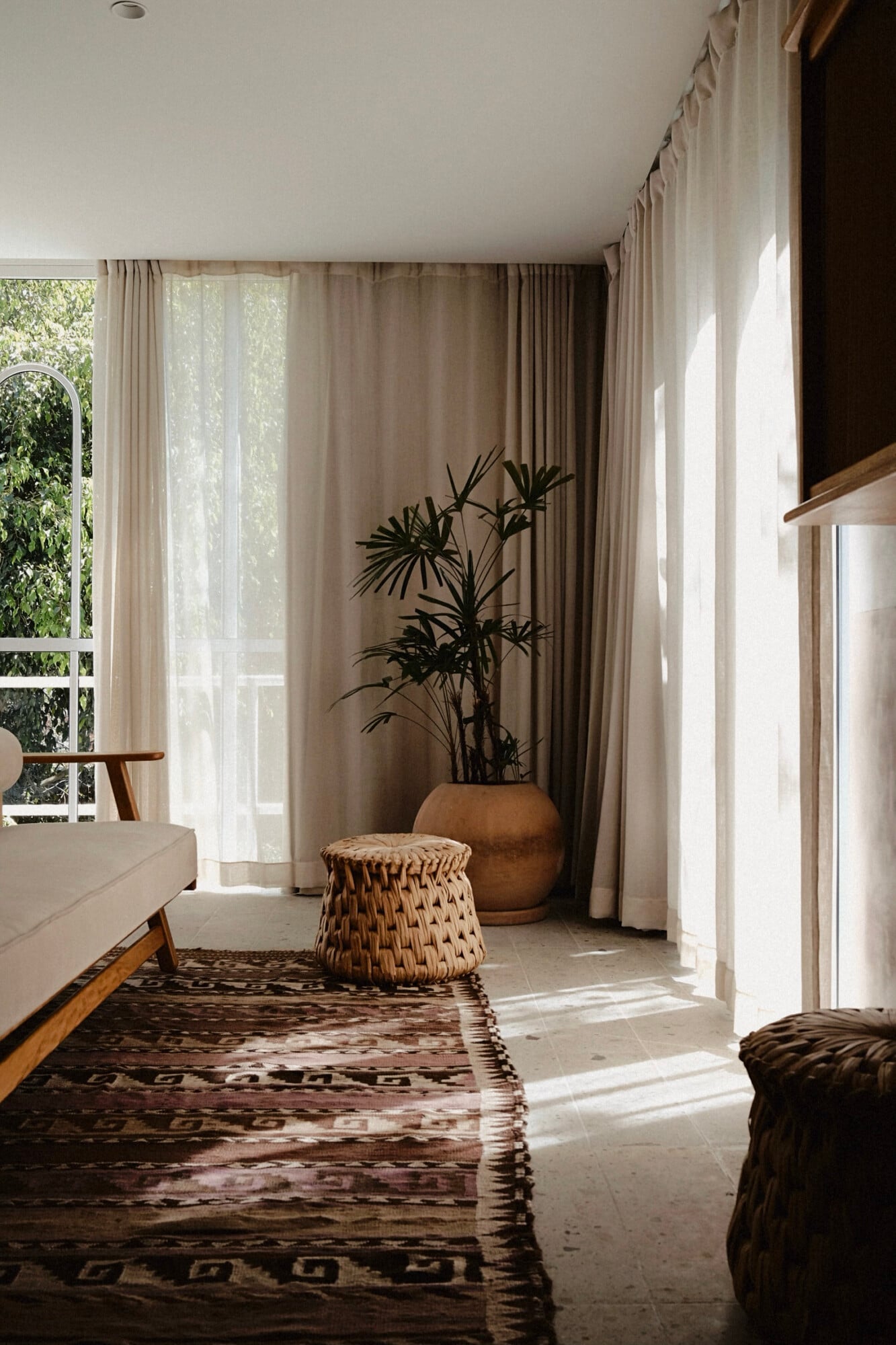 UMA Casa, Mexico — Sunlit bedroom with cream linen sofa, woven poufs, terracotta planter with dark foliage, floor-to-ceiling curtains, geometric patterned runner, and floor-to-ceiling windows overlooking garden.