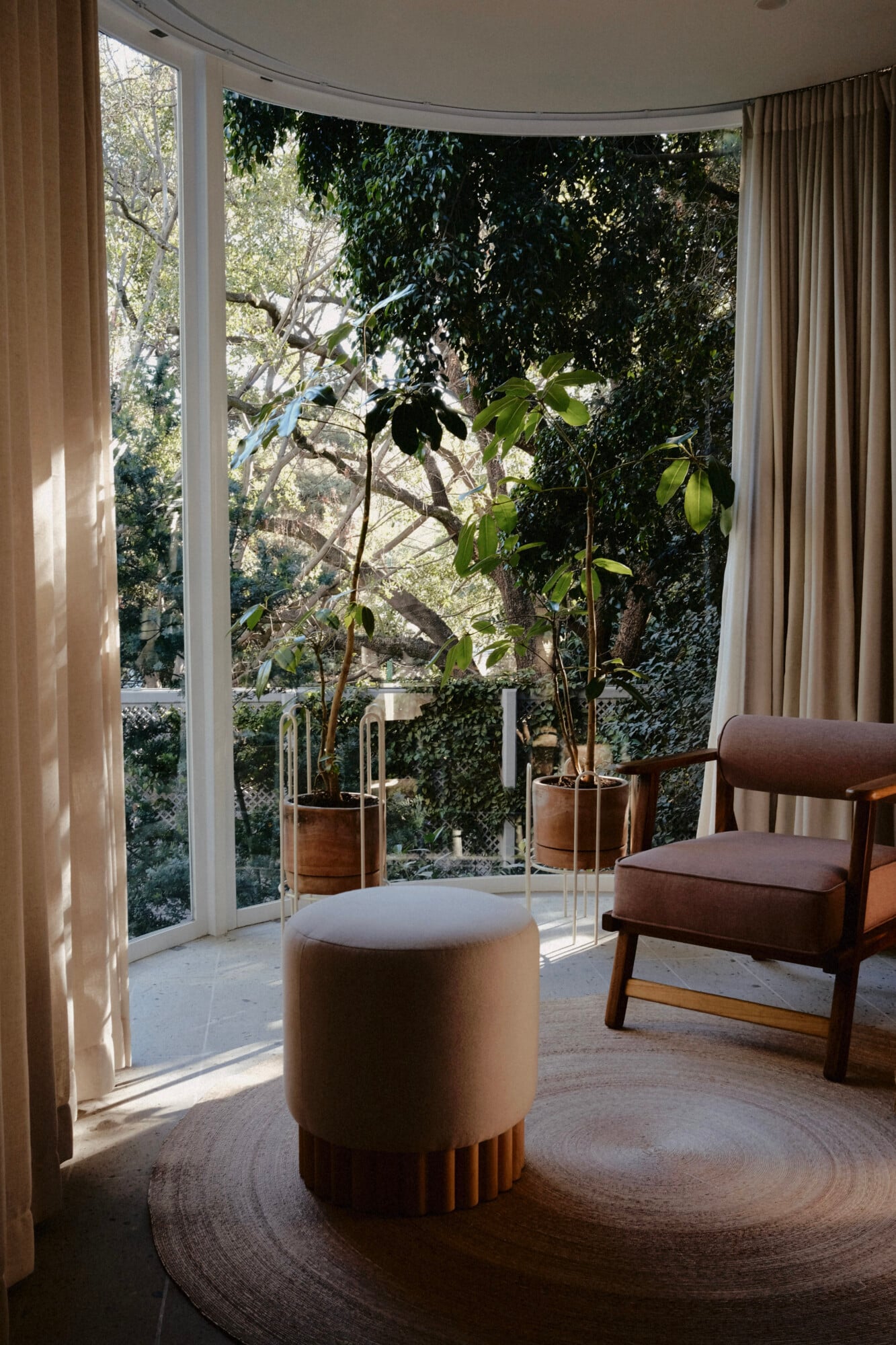 UMA Casa, Mexico — Curved glass doors open to lush gardens from modernist sitting room with natural linen ottoman, mid-century wood chair, and potted trees in dappled sunlight.