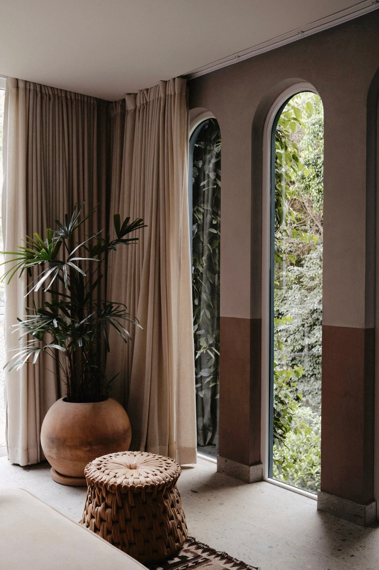 UMA Casa, Mexico — Arched windows frame lush greenery beside taupe curtains, terracotta planter, and woven stool in minimalist interior.