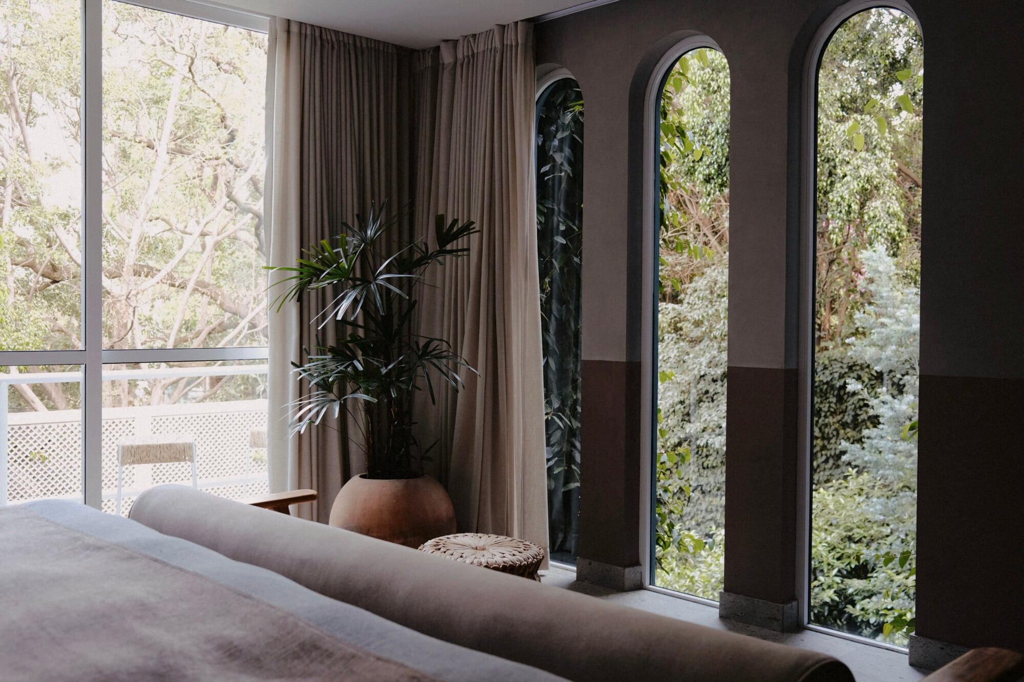 UMA Casa, Mexico — Modern bedroom with floor-to-ceiling arched windows, taupe linen sofa, dark walls, potted plant, and views of lush vegetation through gallery windows.