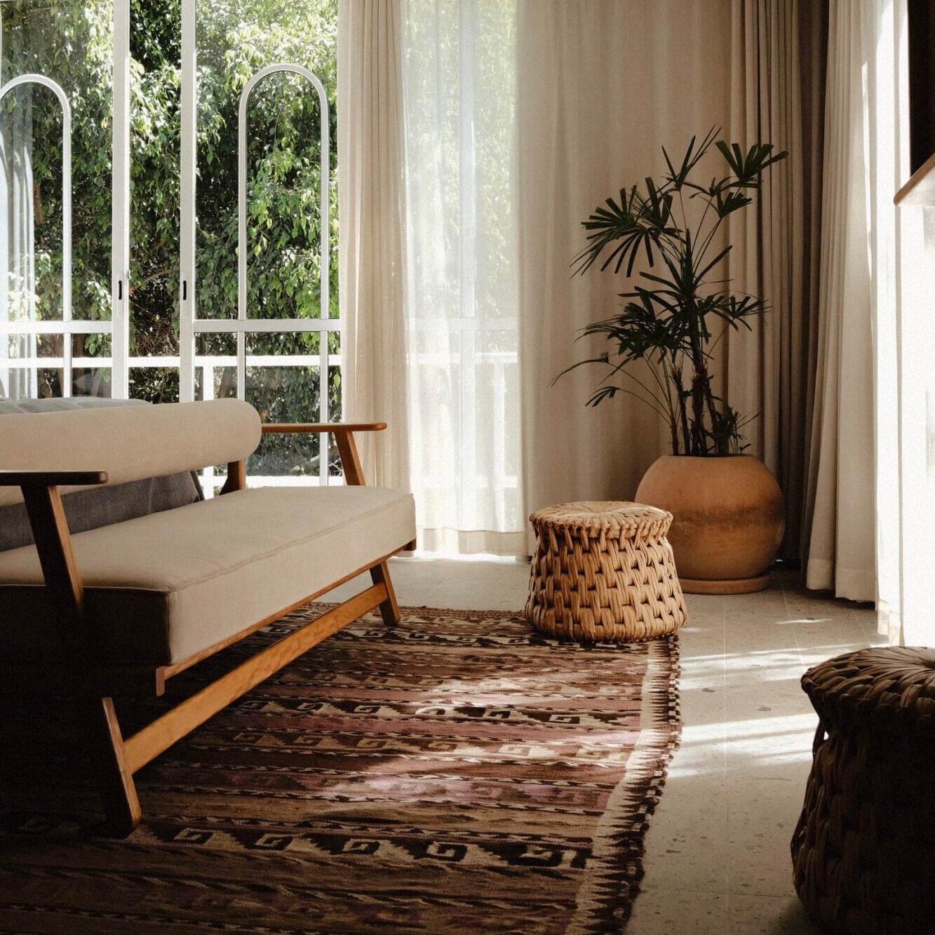 UMA Casa, Mexico — Mid-century cream sofa with wooden frame beside woven basket and terracotta planter with bamboo, arched windows framing garden, natural light through sheer curtains, geometric rug.