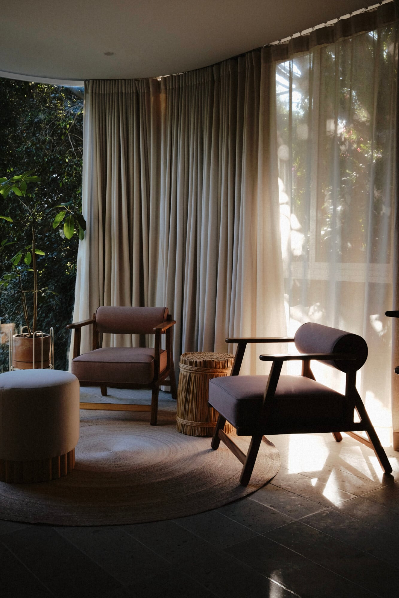 UMA Casa, Mexico — Mid-century modern lounge with dark wood chairs, linen upholstery, woven side table, cream curtains filtering sunlight through floor-to-ceiling windows overlooking garden.
