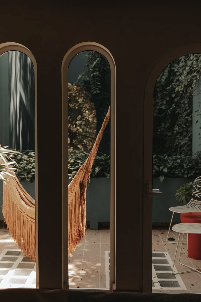 UMA Casa, Mexico — Arched glass doors with bronze frames opening onto courtyard with hammock, ivy-covered walls, and modernist furniture in dappled light.