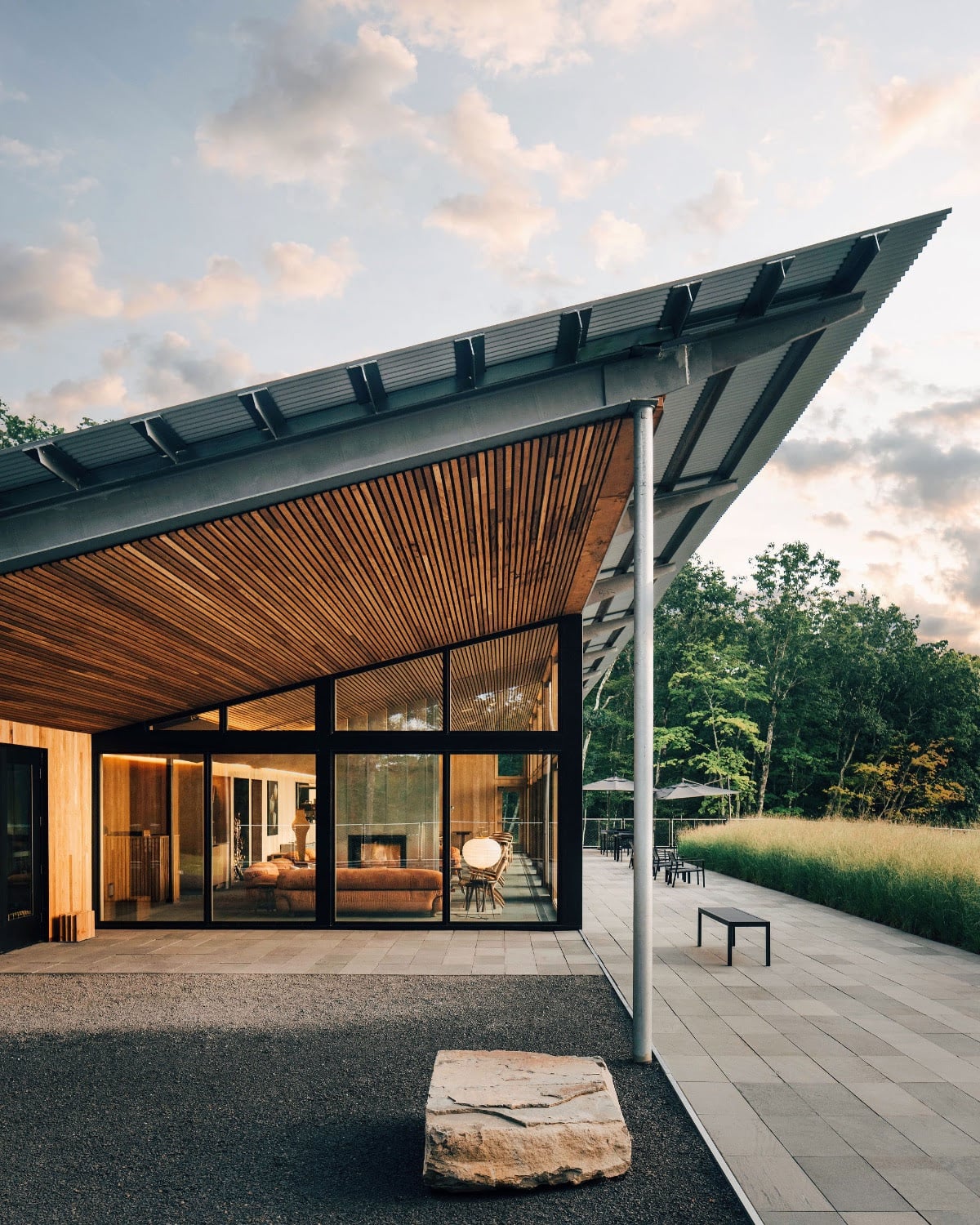 Piaule, North America — Modern pavilion with charcoal metal roof, warm wood interior, floor-to-ceiling glass, concrete terrace, and forest backdrop at dusk.
