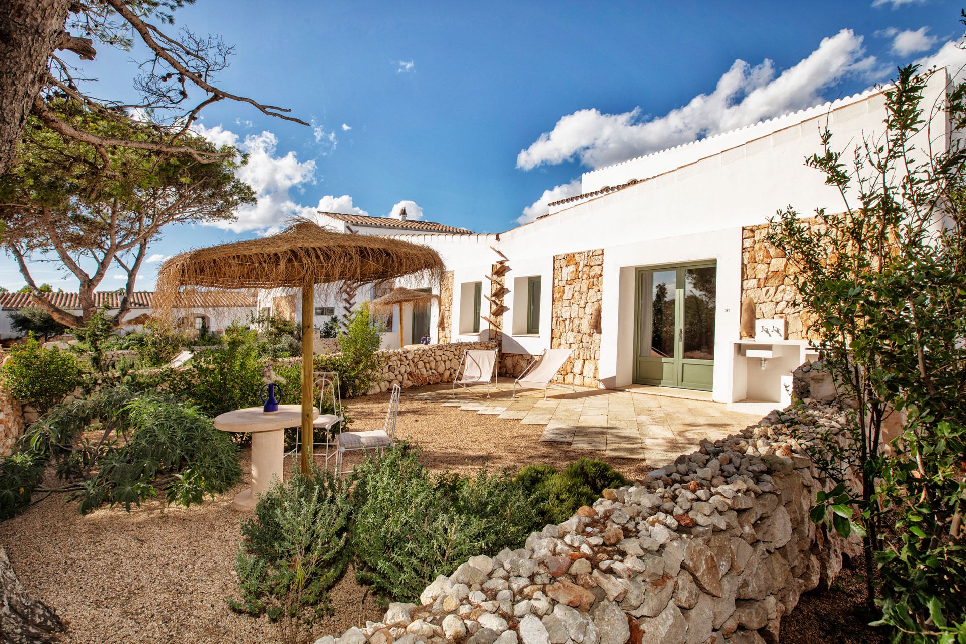 Menorca Experimental, Balearic Islands — Whitewashed Mediterranean villa courtyard with thatched pergola, stone terracing, and clear sky.