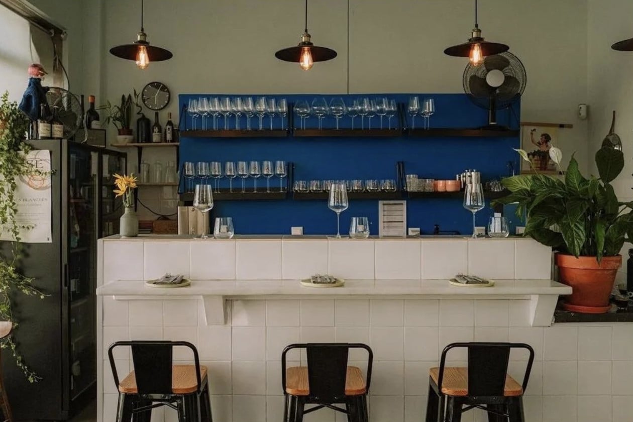 Senhor Uva, Portugal — Modern wine bar with blue-backed shelving, white tile counter, pendant lighting, and black bistro seating.