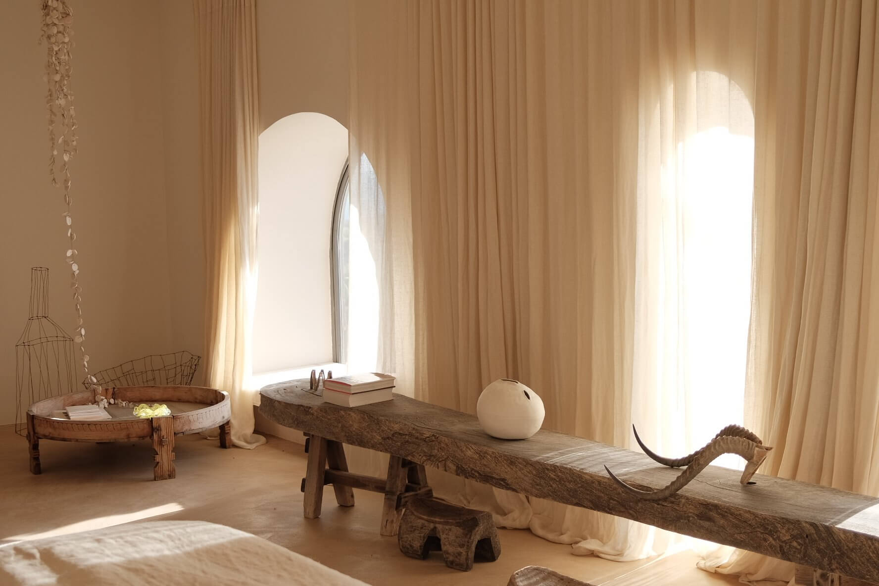 Parco Dei Sesi, Europe — Minimalist wellness room with natural wood beam furniture, arched window, cream curtains, and soft diffused light creating serene spa-like atmosphere.