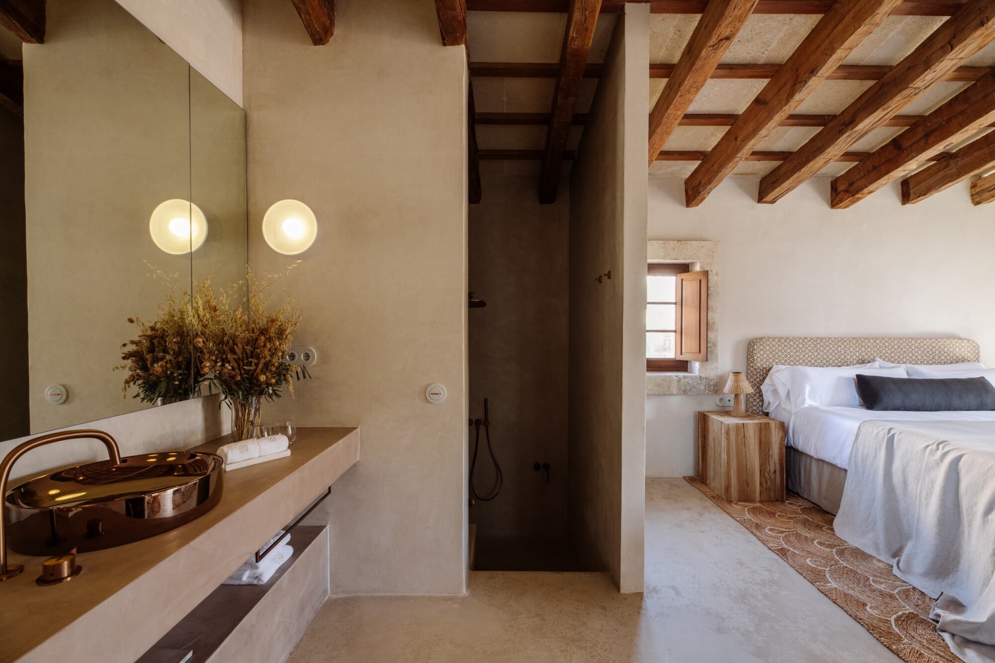 Santa Ana, Balearic Islands — Ensuite bathroom with brass sink, mirrored vanity, exposed wooden beams, and open passage to bedroom with neutral linens and natural light.