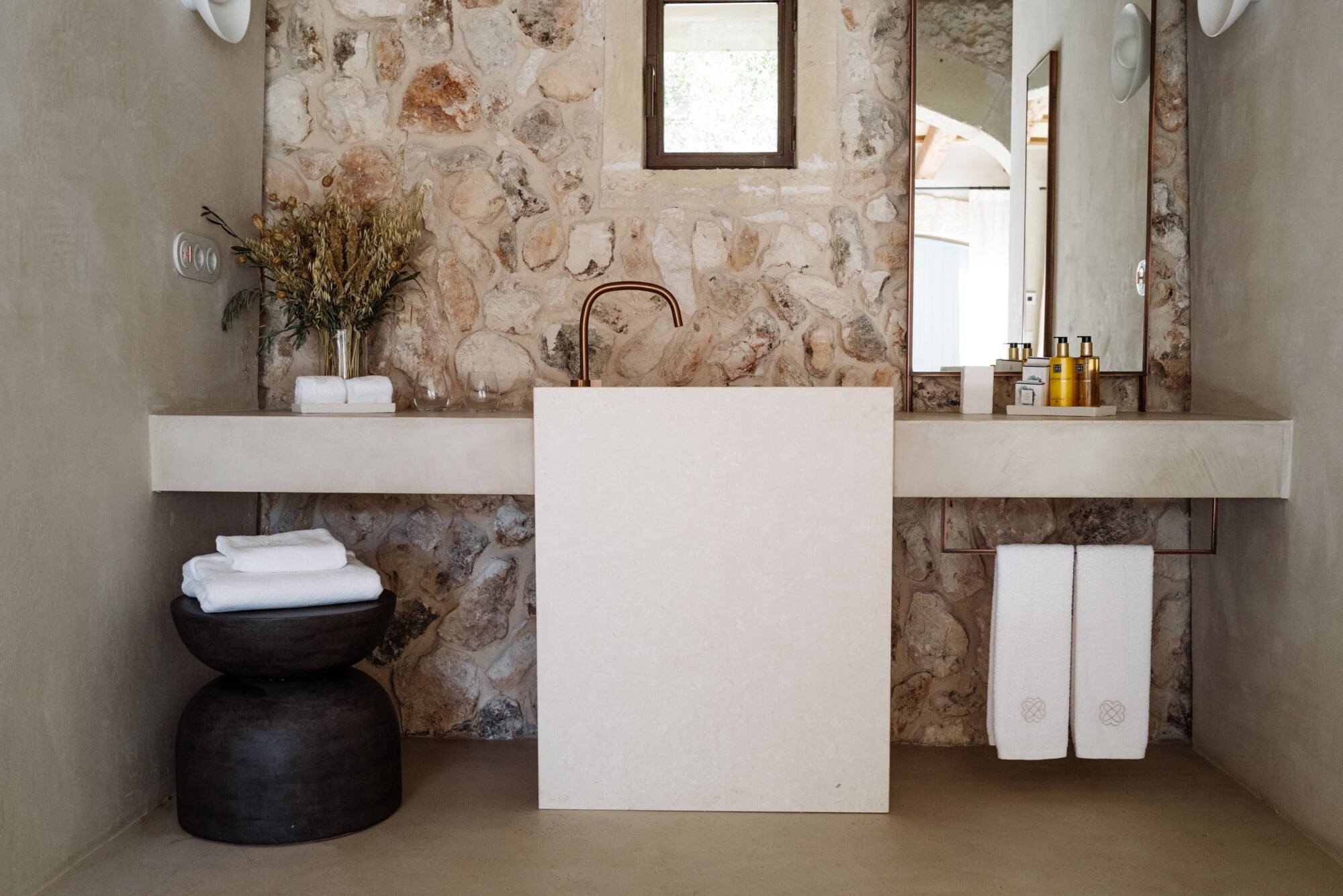 Santa Ana, Balearic Islands — Minimalist bathroom with travertine stone wall, dual floating vanities, white vessel sink, and black ceramic towel vessel.