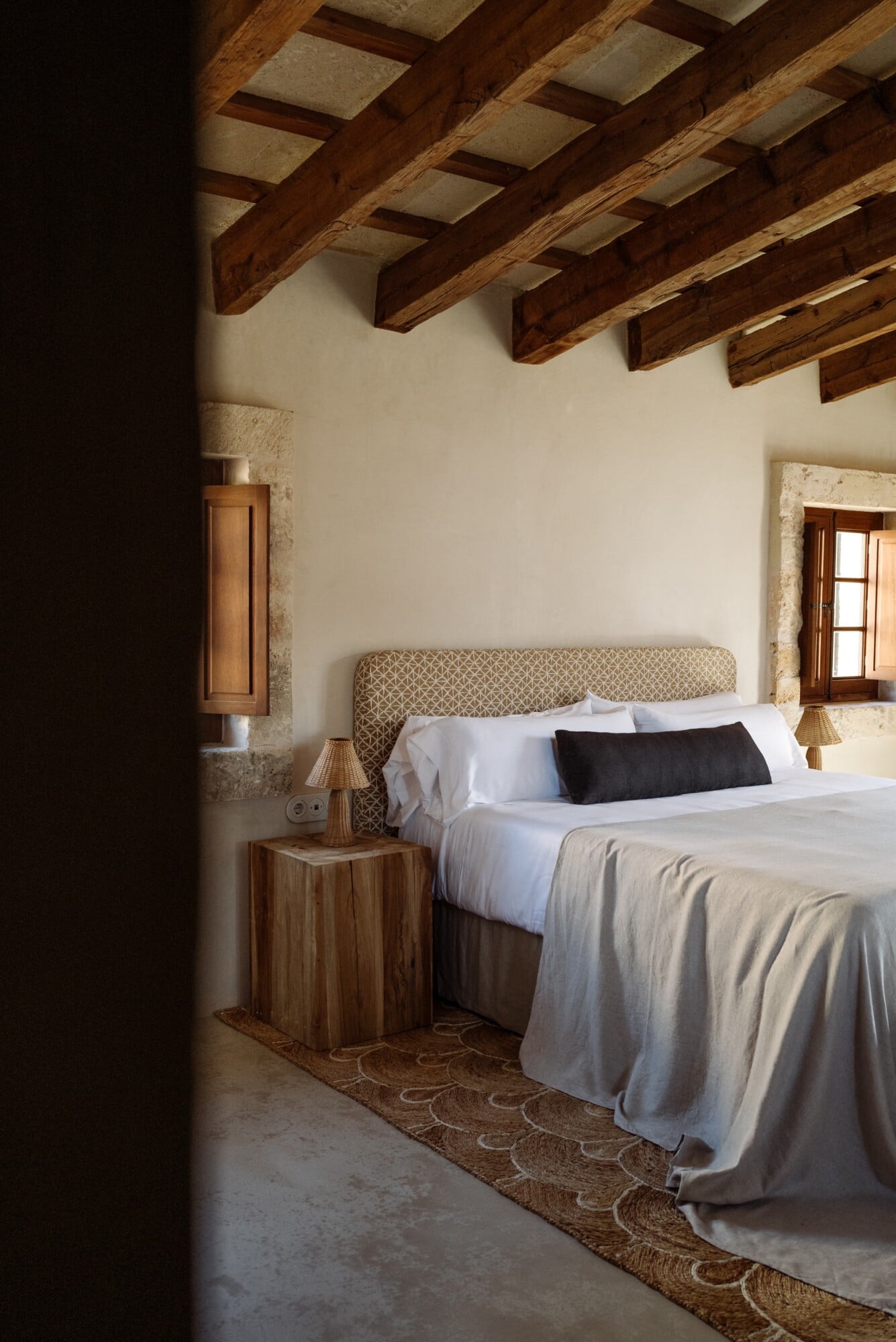 Santa Ana, Balearic Islands — Bedroom with exposed wooden beams, stone walls, patterned headboard, natural light, neutral linens, and woven area rug.