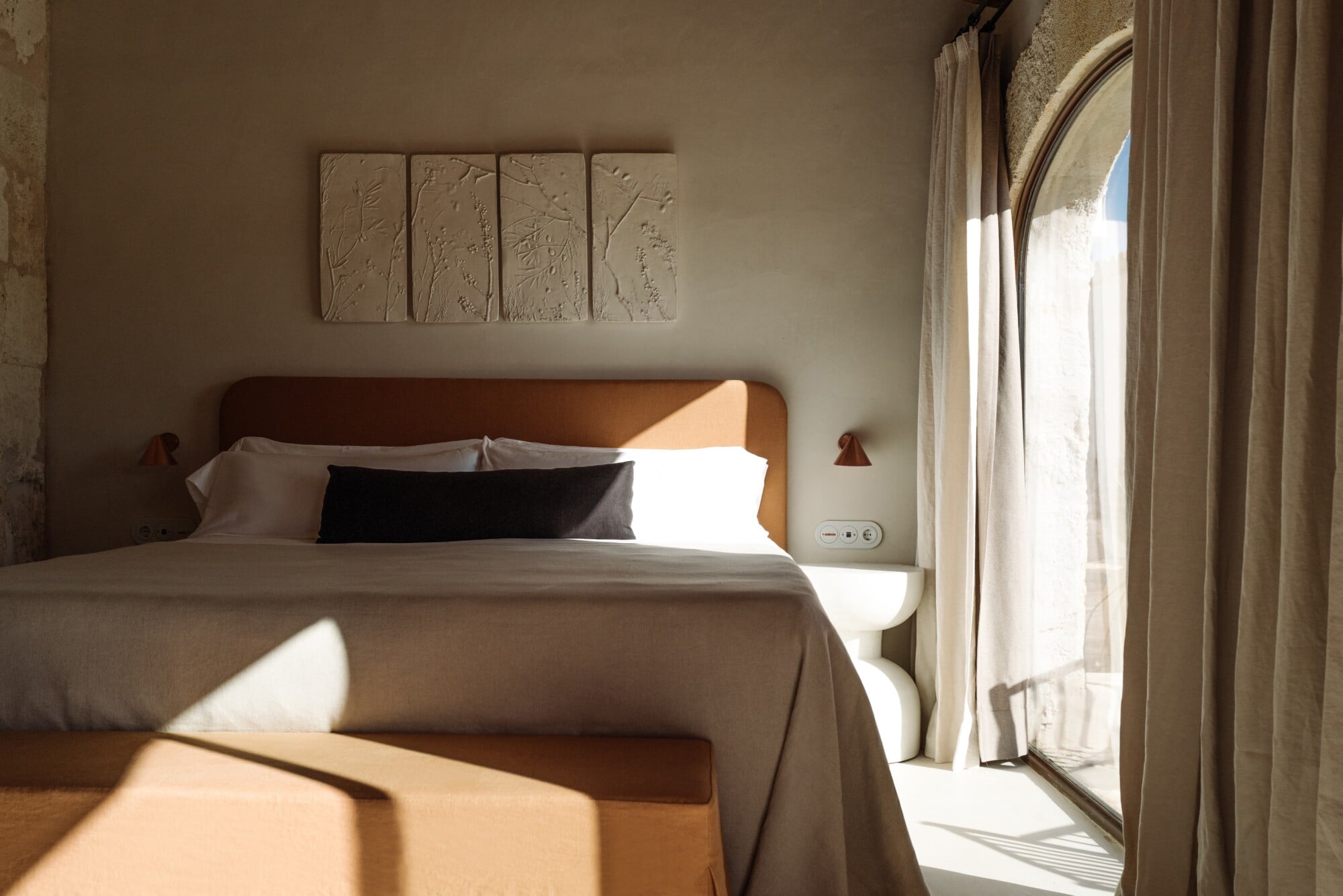 Santa Ana, Balearic Islands — Minimalist bedroom with wooden headboard, linen bedding, textured wall panels, arched doorway, and golden afternoon light through sheer curtains.