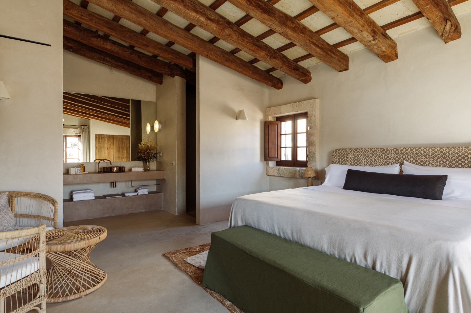 Santa Ana, Balearic Islands — Bedroom with exposed timber beams, stone sink vanity, woven rattan chairs, green bench, and wooden shuttered window.