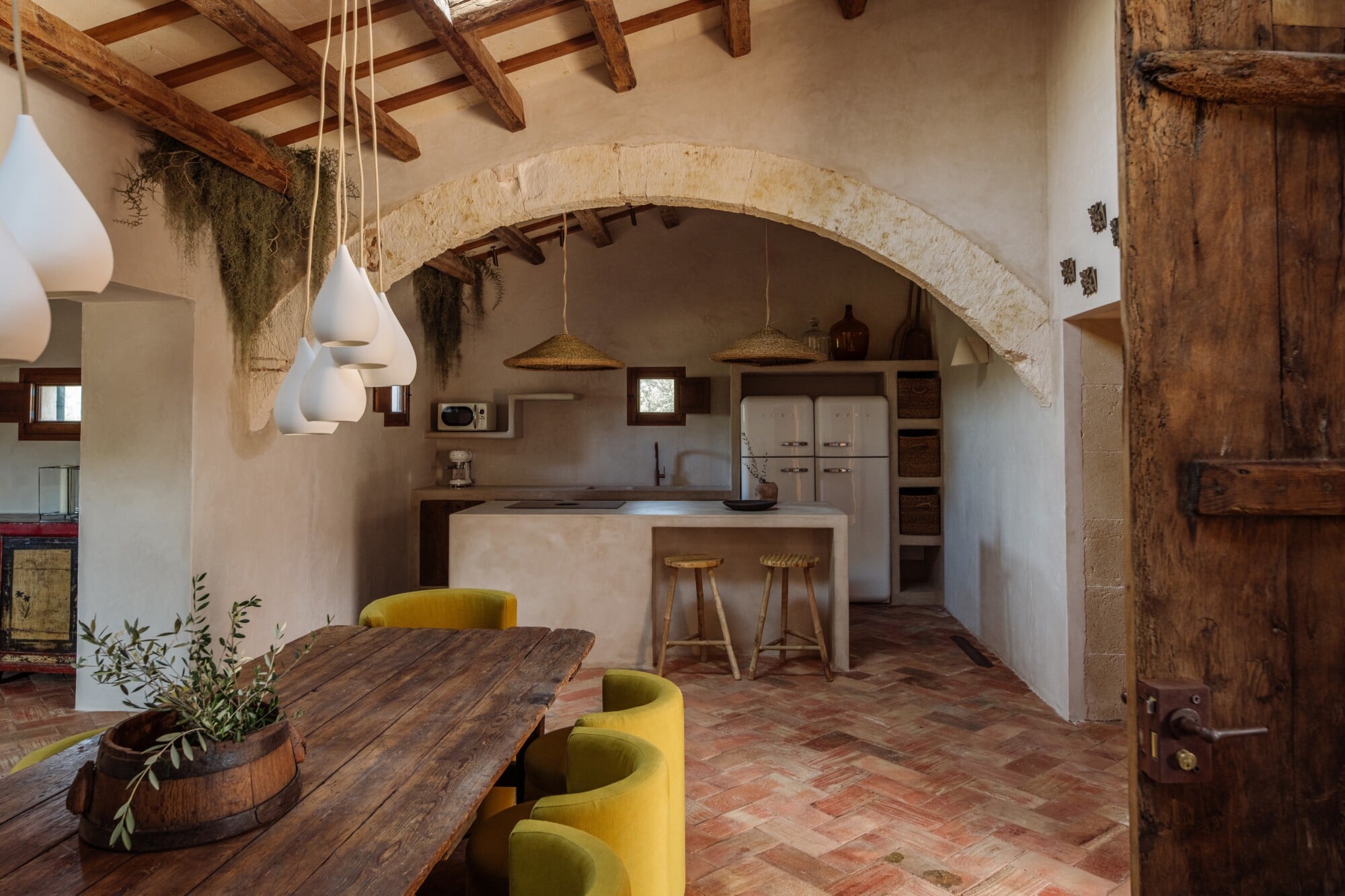 Santa Ana, Balearic Islands — Arched stone kitchen with exposed timber beams, yellow chairs, woven pendant lights, terracotta floors, rustic wood details.